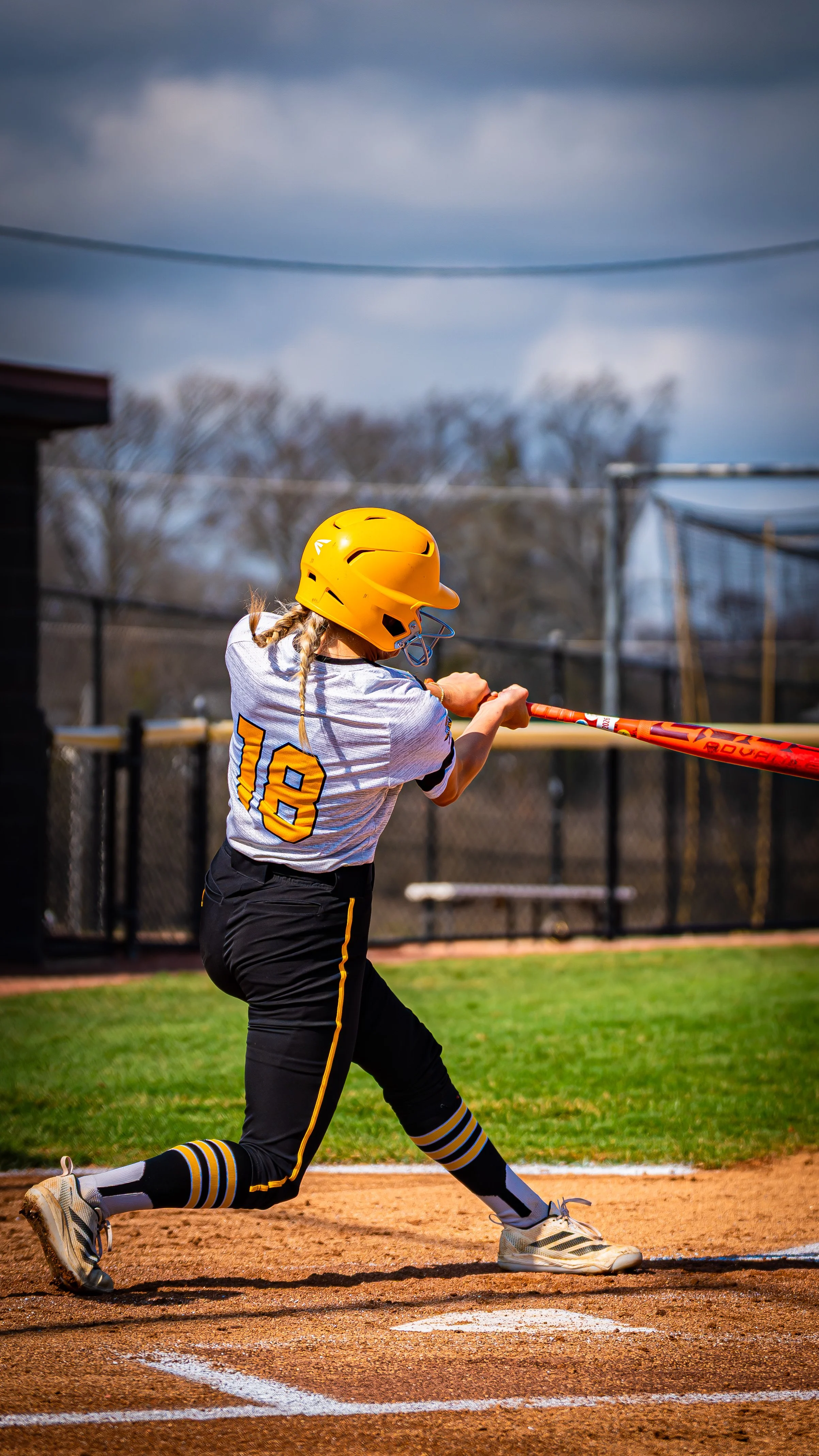 Female softball player wearing a yellow helmet and a white jersey with the number 18, swinging a red bat at a ball on the field.