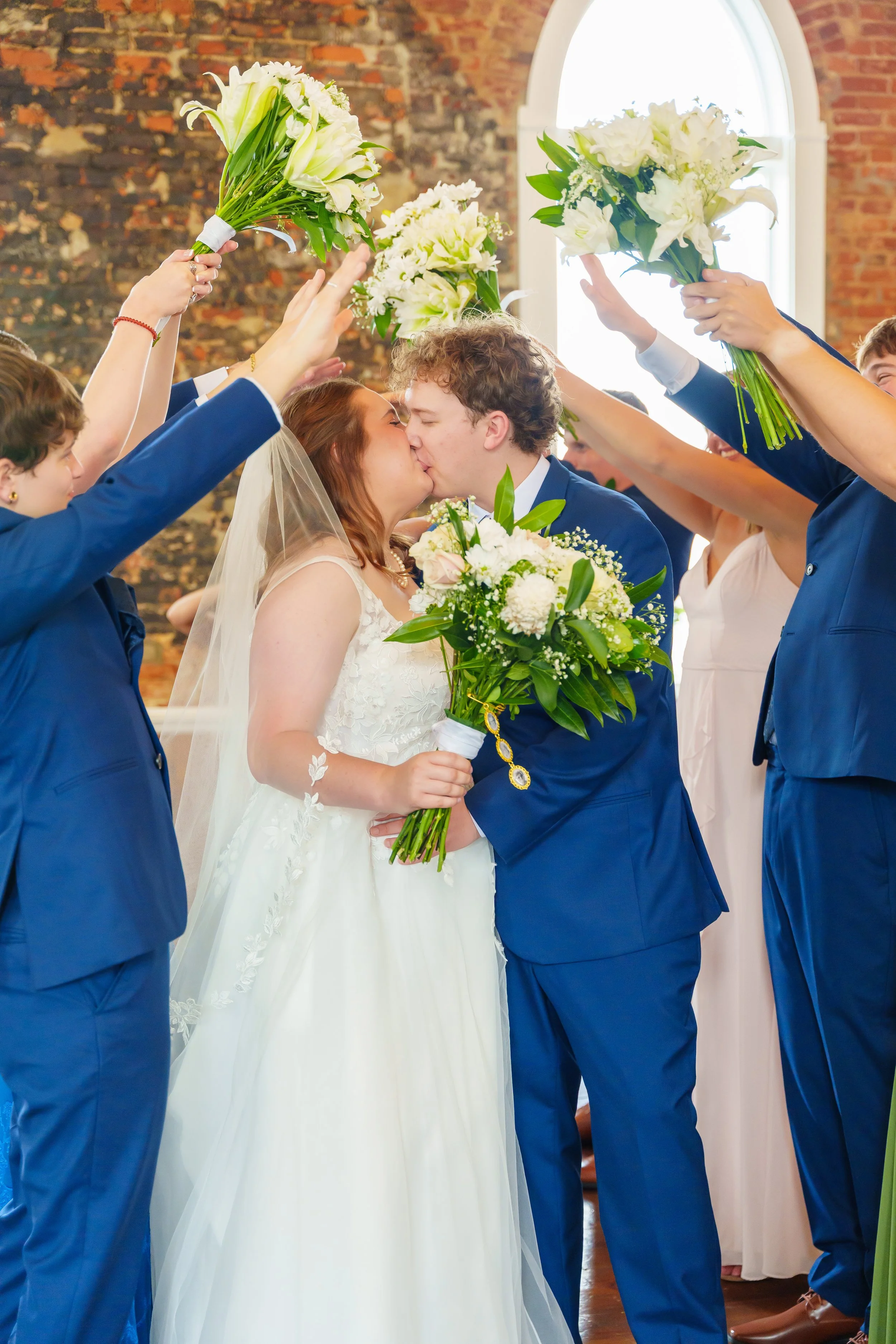 A bride and groom kiss during their wedding ceremony, surrounded by friends holding bouquets of white flowers raised in celebration inside a brick-walled venue with a large arched window.