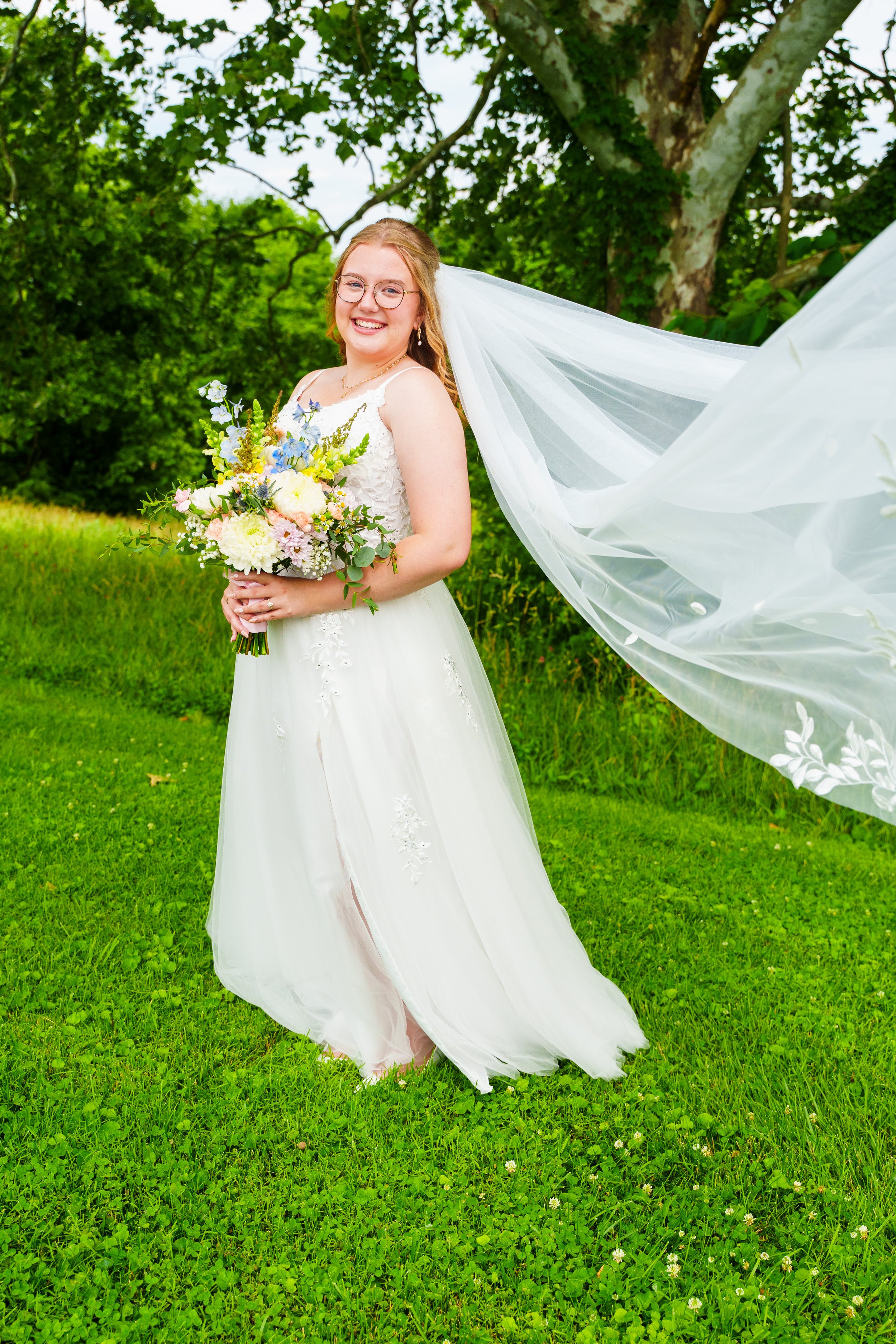 A smiling bride in a white wedding dress holding a bouquet of flowers outdoors, with a flowing veil and green trees in the background.