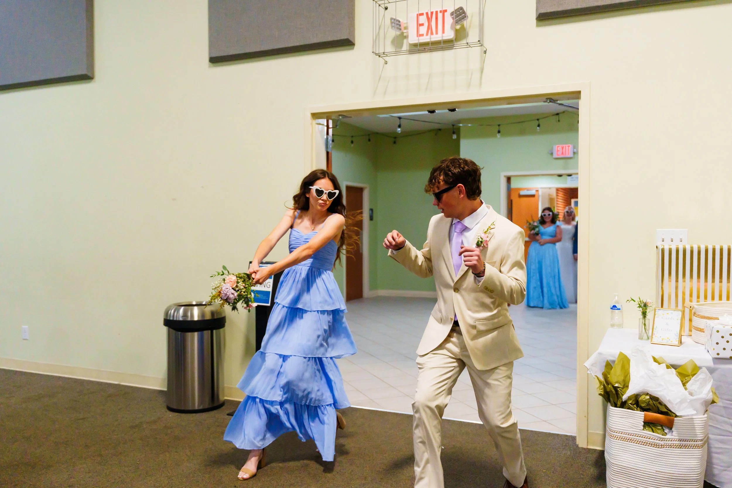 A woman in a blue dress and white heart-shaped sunglasses dances with a man in a beige suit at a celebration or wedding reception. Two women in blue dresses stand in the background near a hallway. Decor includes flowers, a sign, and a table with item