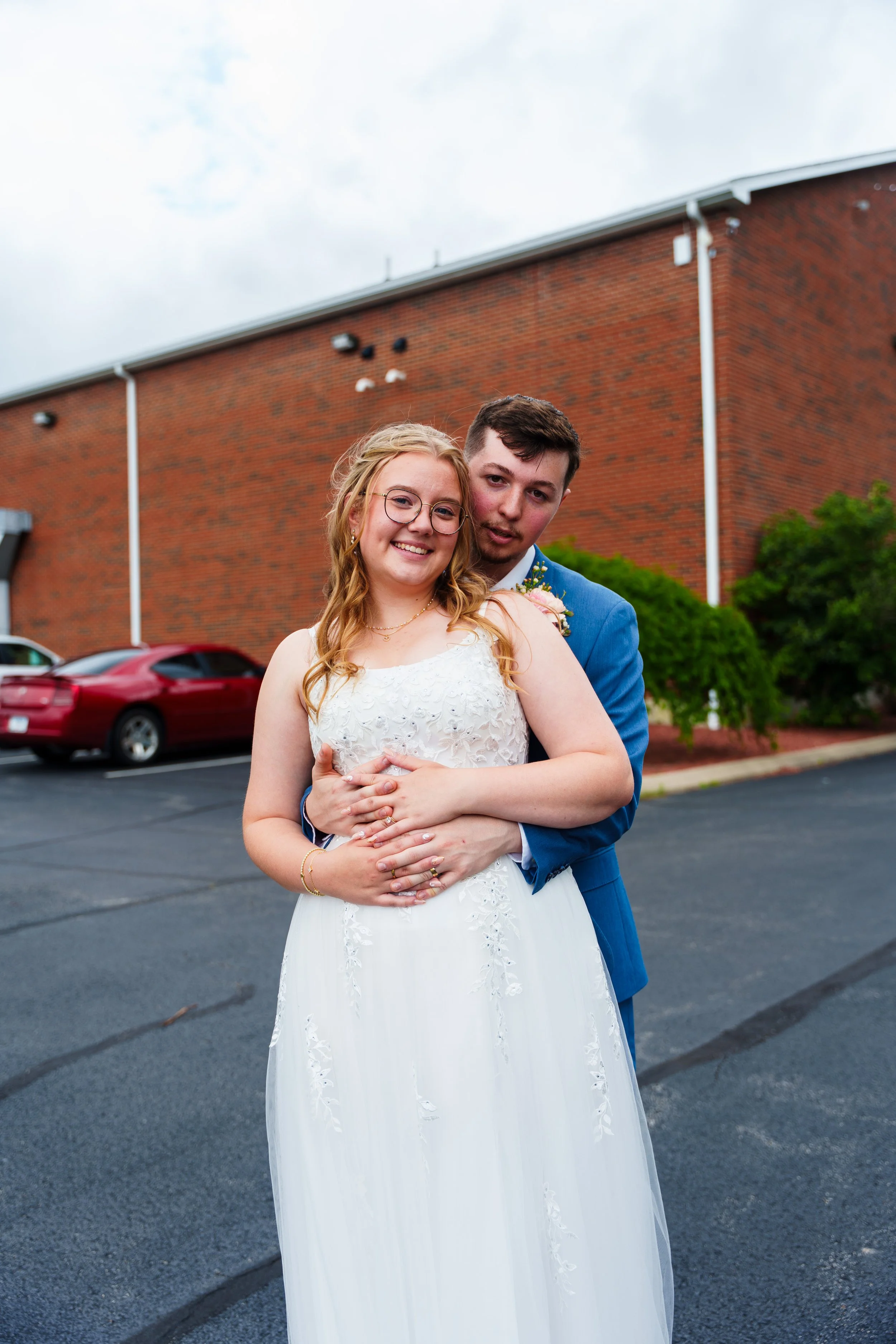 A bride and groom standing outdoors in front of a brick building, smiling and hugging, on a cloudy day.
