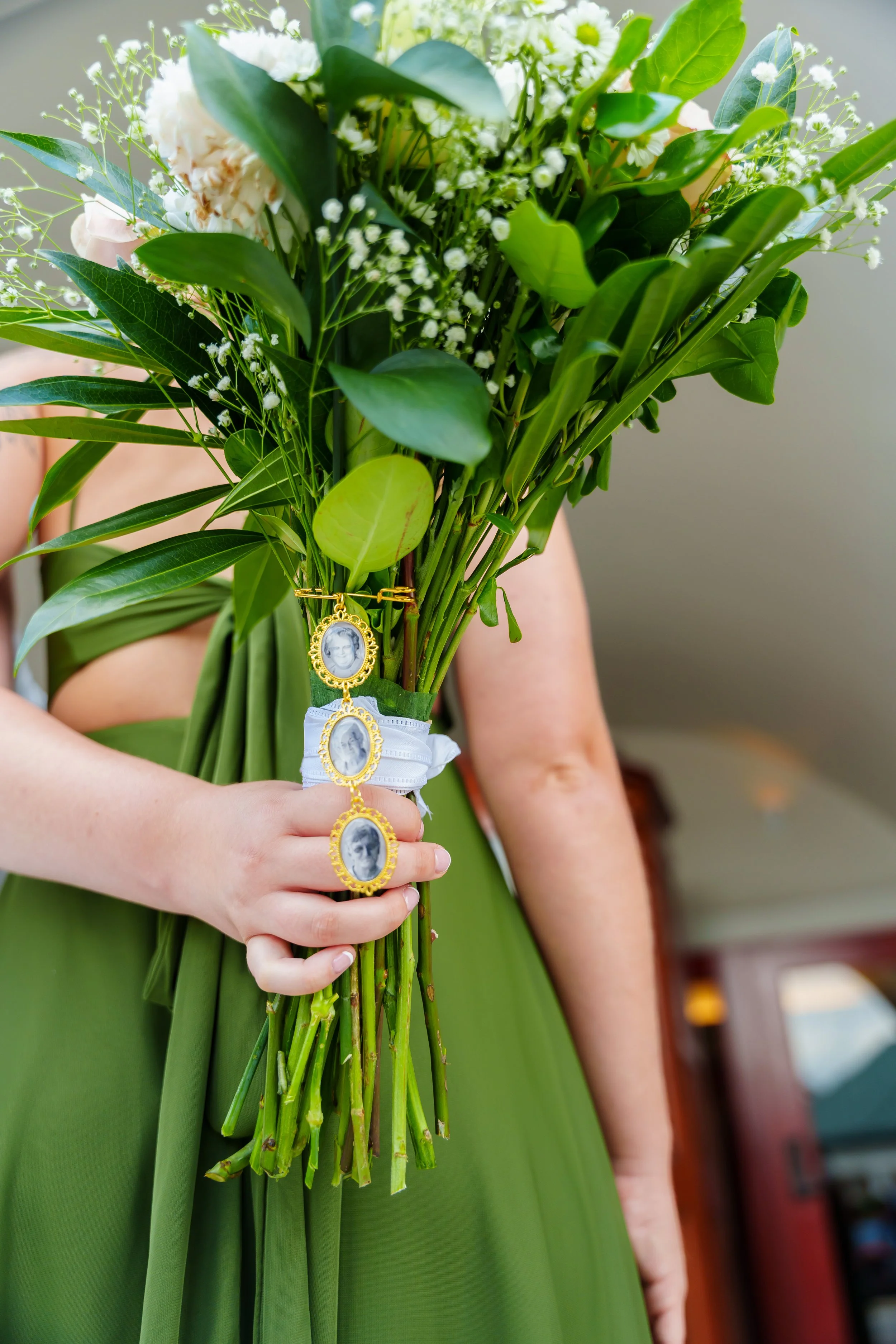 A person in a green dress is holding a bouquet of white flowers with greenery and a small charm with a black-and-white photo of a woman.