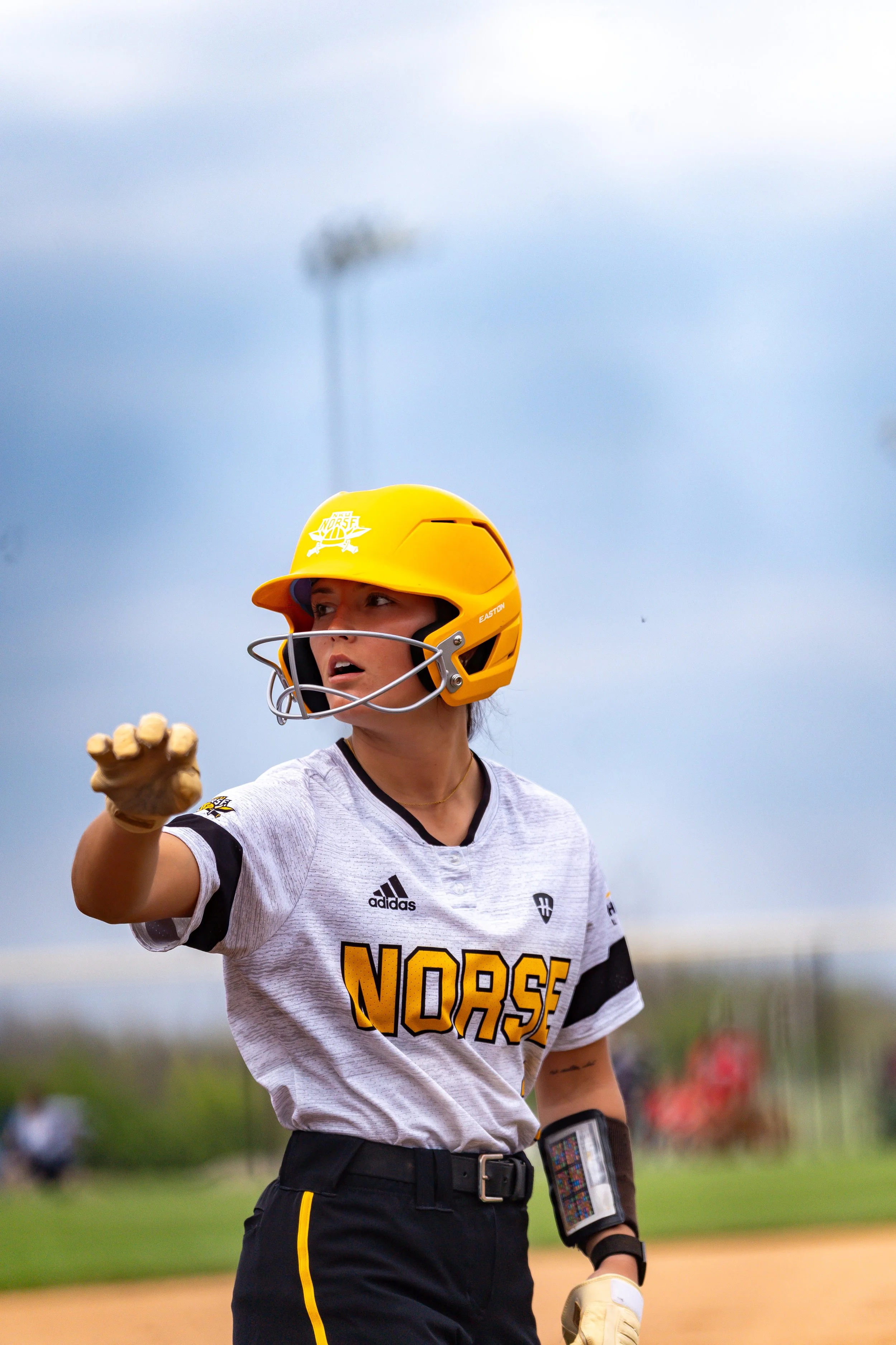 Female softball player in a white jersey with black and yellow details, wearing a yellow helmet, on a baseball field with cloudy sky in the background.