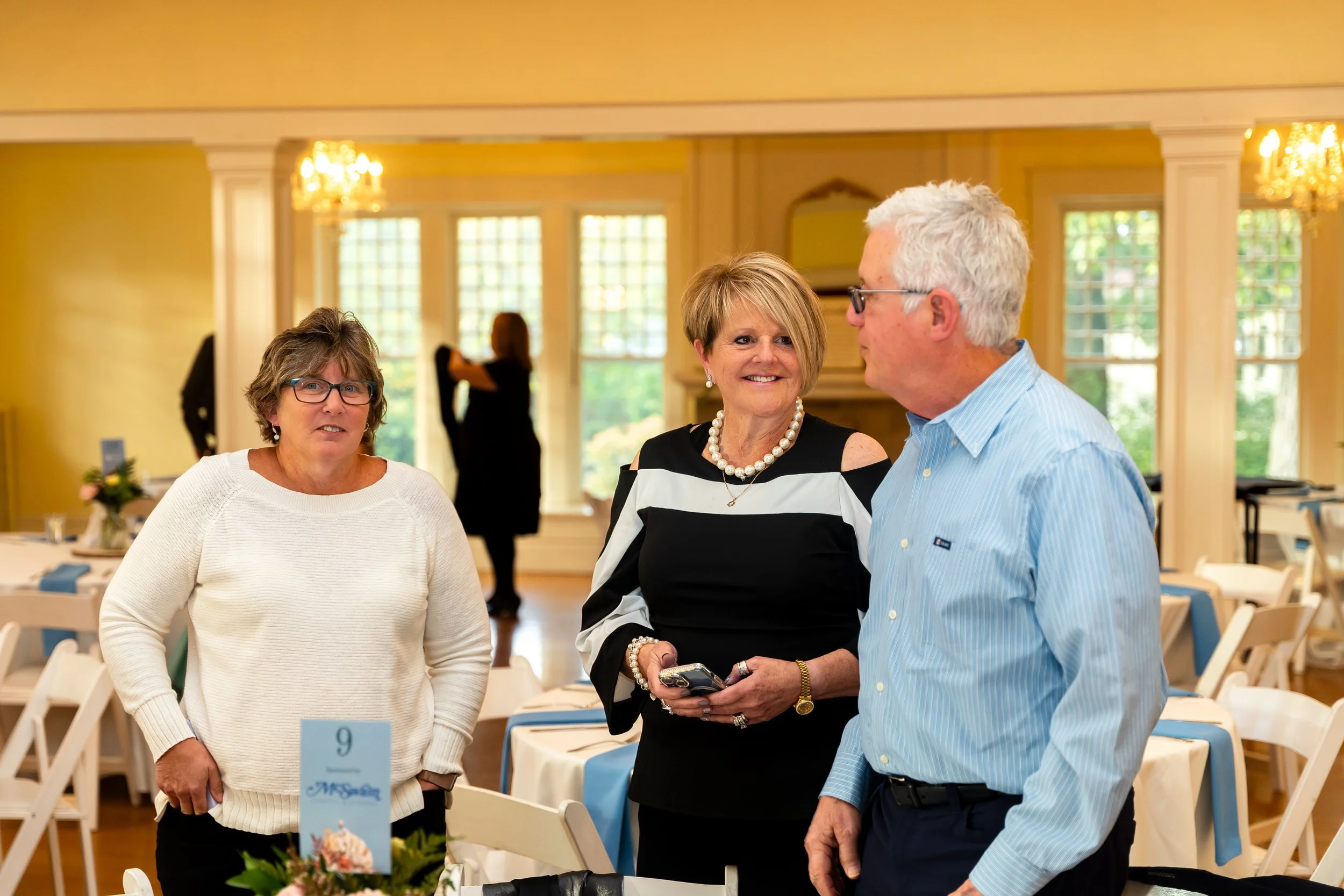 Three middle-aged adults, two women and one man, are engaging in conversation at a social event in a warmly-lit, elegant room with large windows and chandeliers. They are standing near tables decorated with flowers.