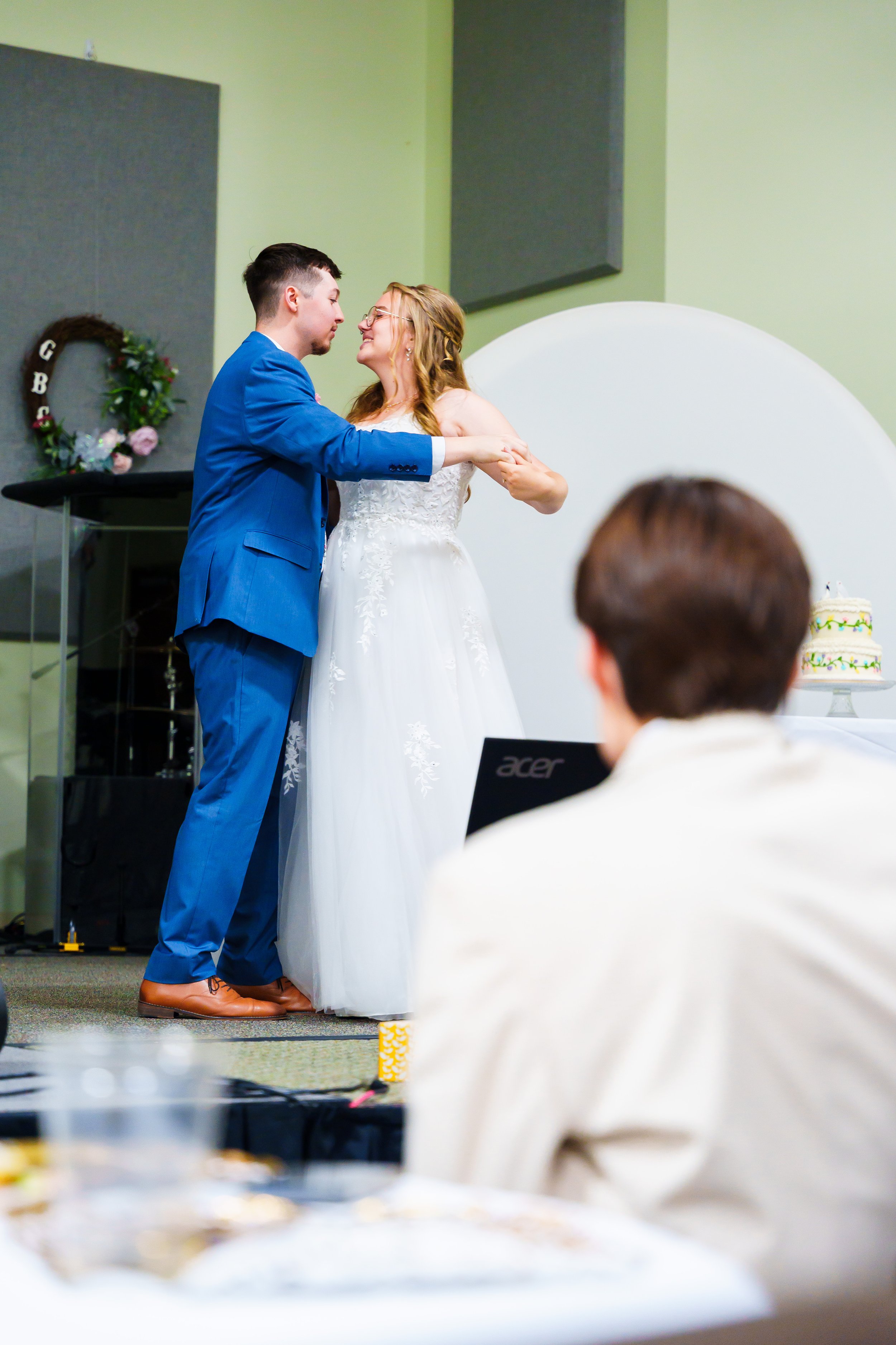 A couple is dancing closely at a wedding reception; the man in a bright blue suit and the woman in a white wedding gown are smiling and holding hands.