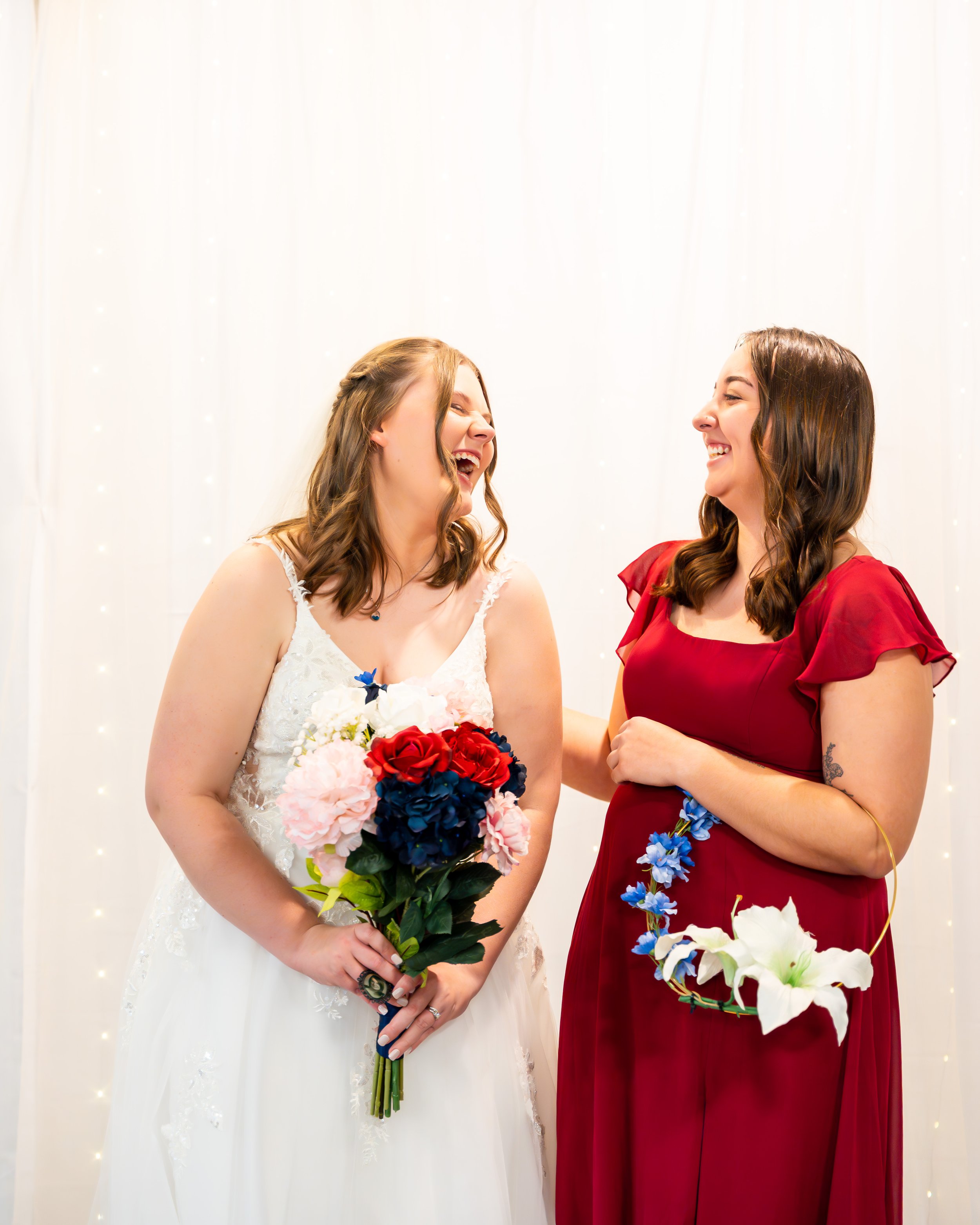 Two women in wedding attire smiling and holding flowers, standing against a white backdrop.
