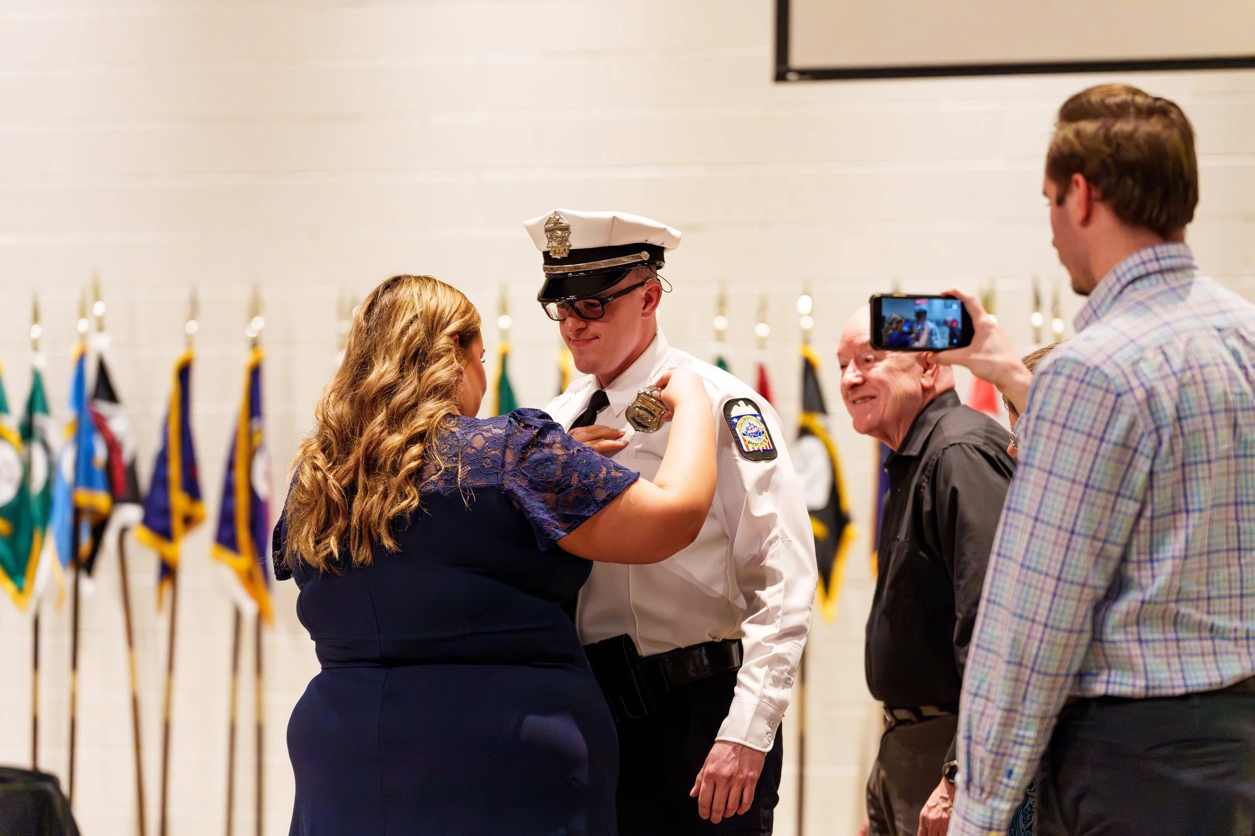 A woman and a police officer share a dance during a ceremony, with a man taking a photo of them and another man smiling nearby. Background features a row of flags.