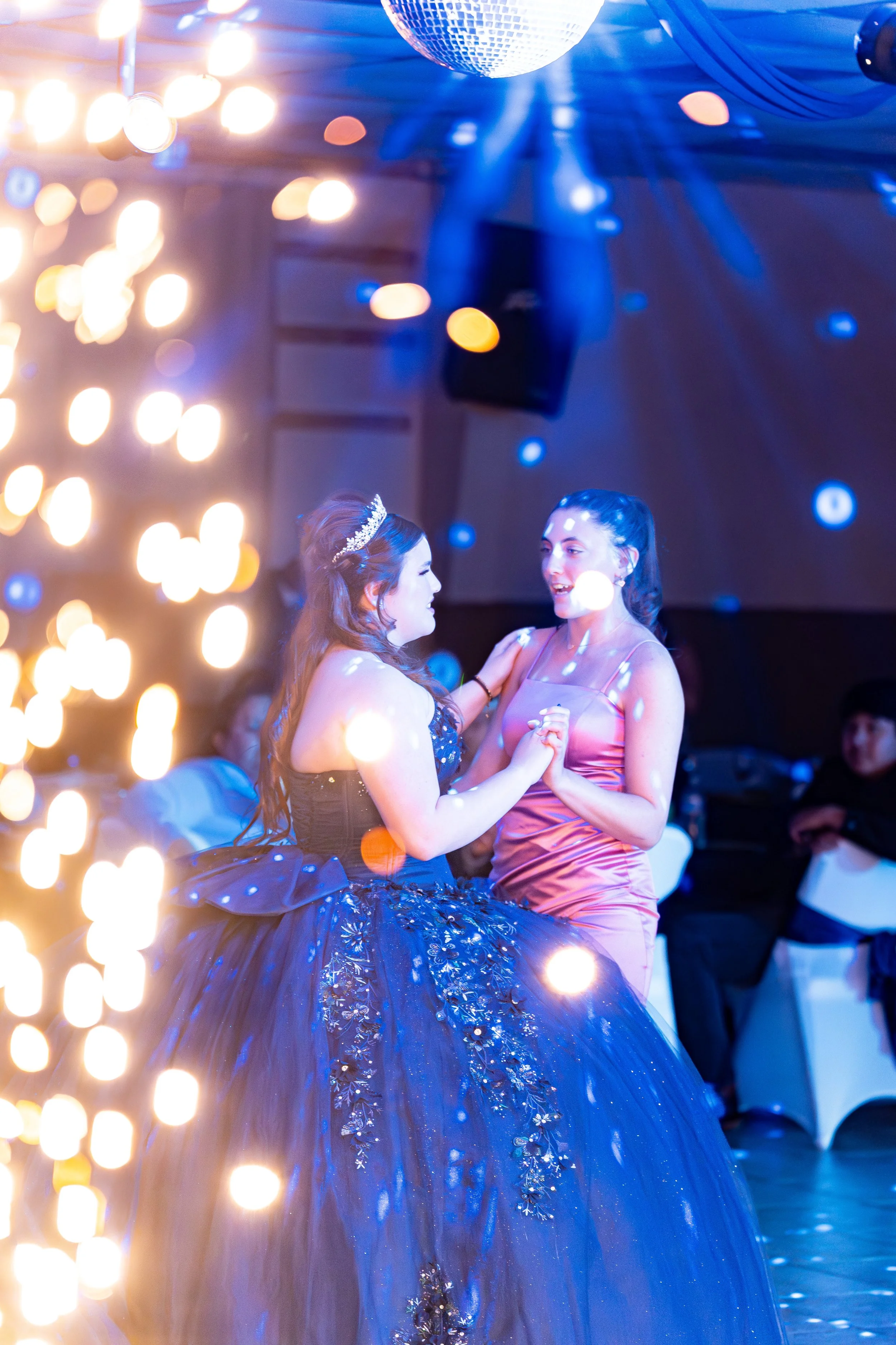 Two women dancing at a party or celebration, one wearing a tiara and a dark blue gown with floral details, and the other in a pink satin dress. There are colorful lights and disco balls in the background, creating a festive atmosphere.