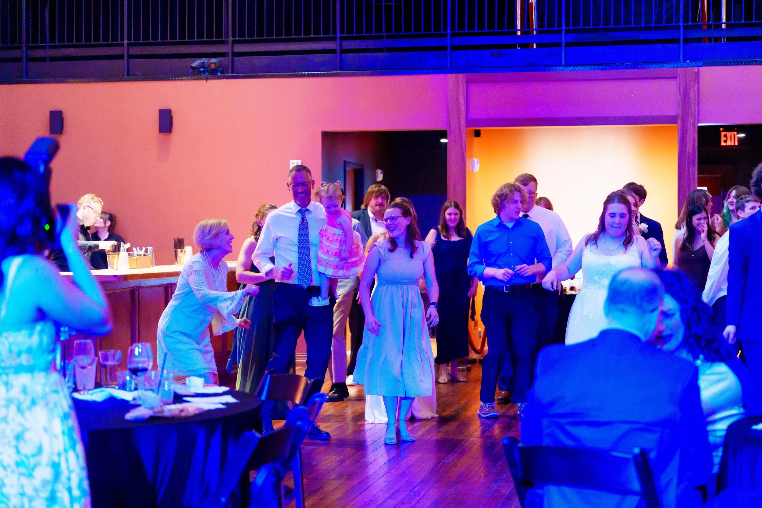 Group of people dancing and socializing at a party or wedding reception in a dimly lit room with colorful lighting.