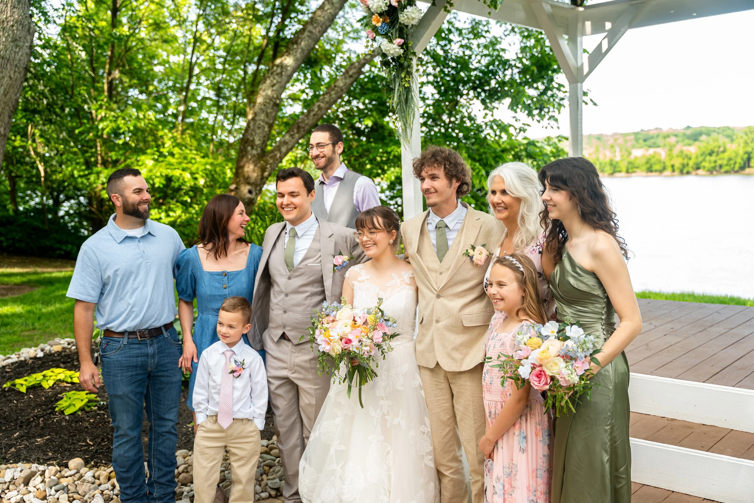 A group of people celebrating a wedding outdoors near a lake with green trees. The bride is holding a bouquet and wearing a white dress, surrounded by friends and family dressed in semi-formal clothes, smiling and happy.