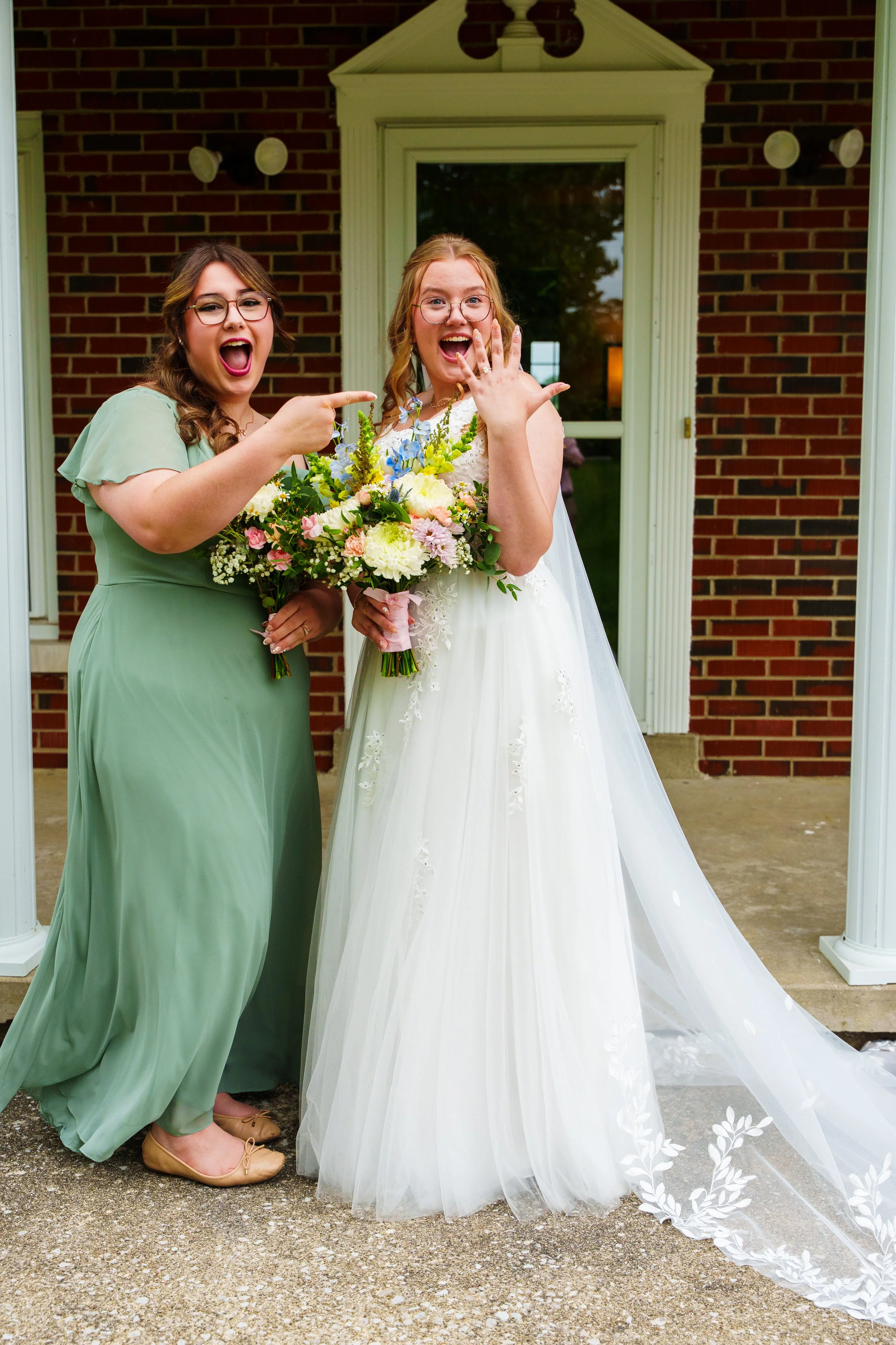 Two women, one in a white wedding dress holding a bouquet of flowers, and the other in a green dress pointing at the bride's left hand which shows a wedding ring, standing in front of a brick house with a porch.