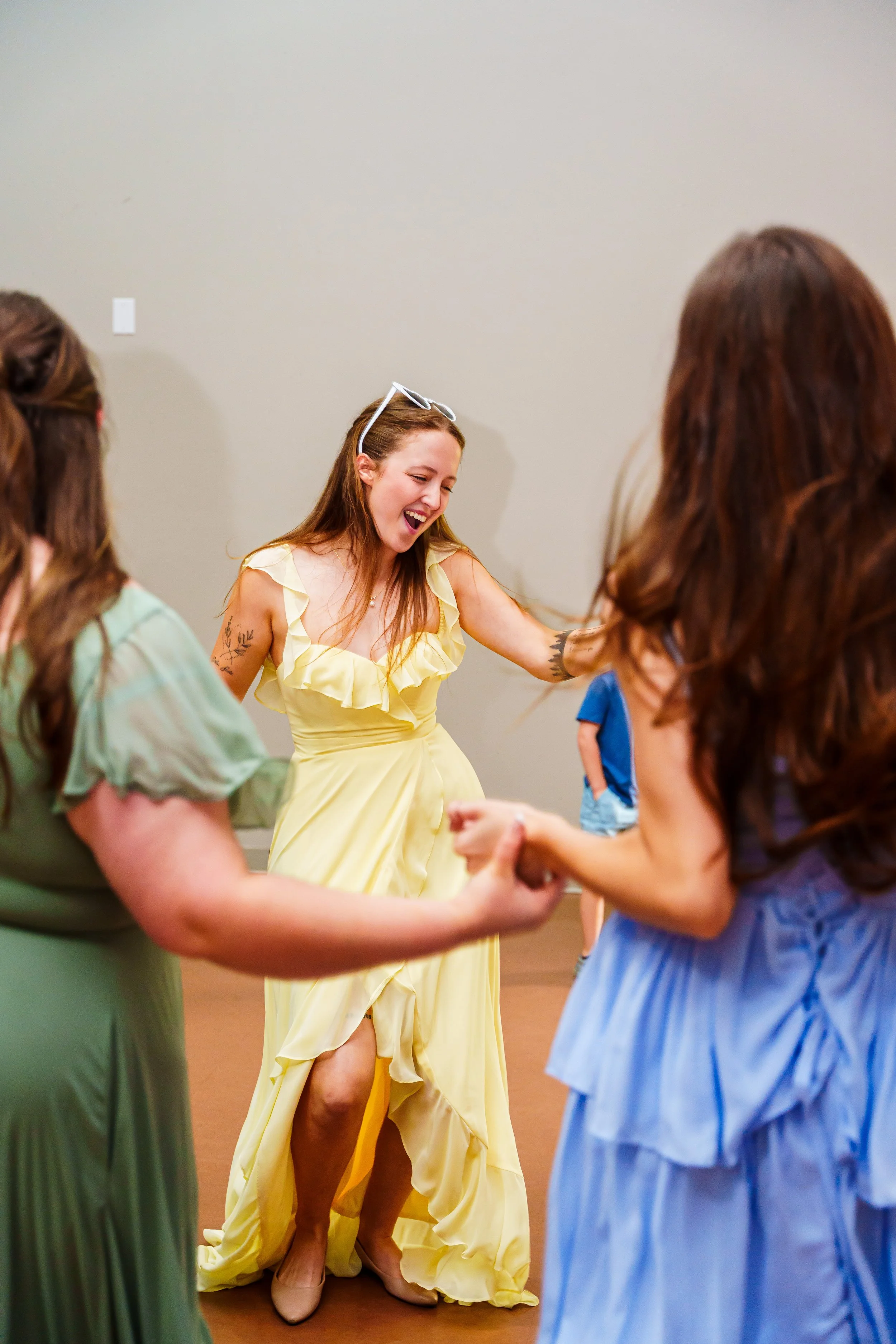 A woman in a yellow dress laughing and dancing with multiple other women in colorful dresses.