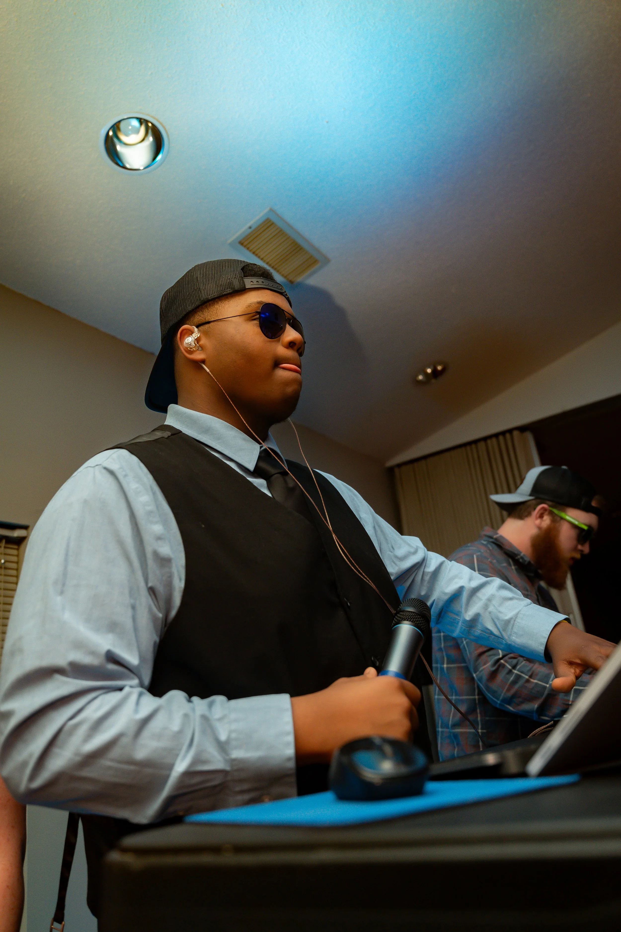 Young man wearing sunglasses, a backward cap, and earbuds, working on a DJ mixer at a party or event.