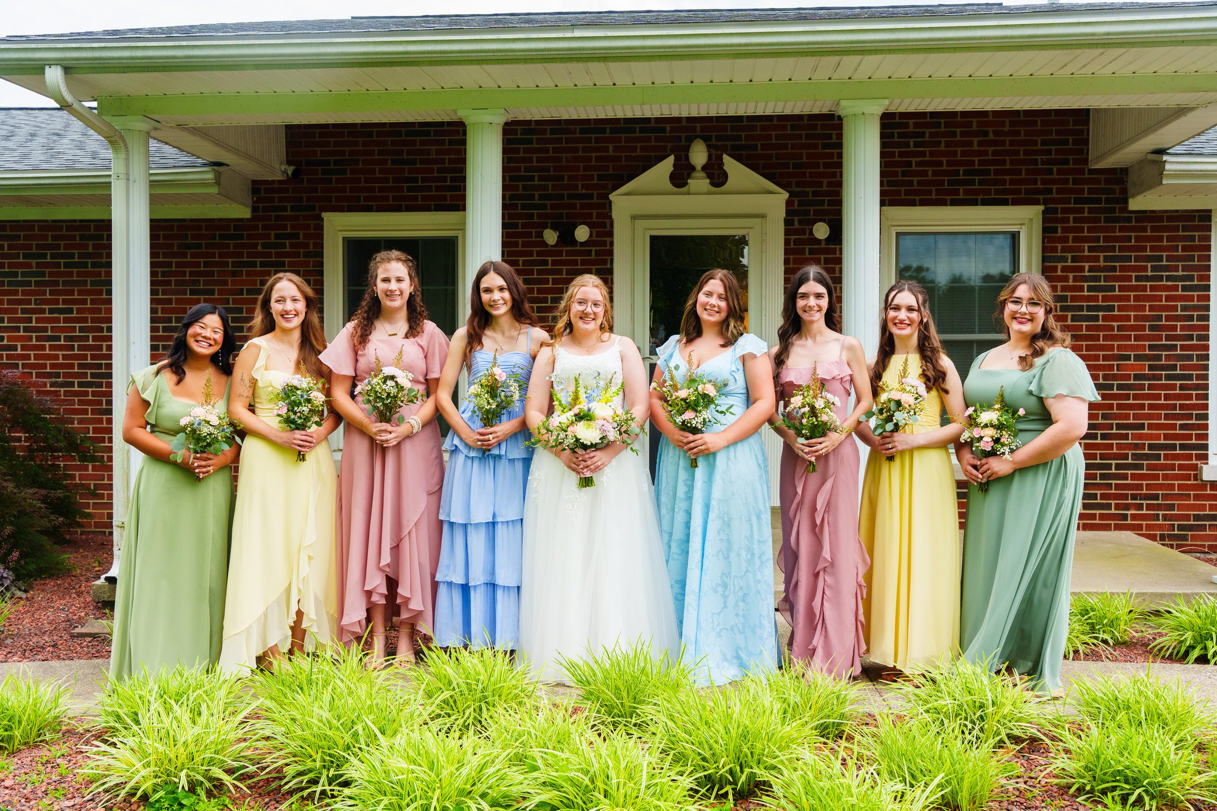 A group of nine young women in pastel-colored dresses holding bouquets of flowers, standing in front of a brick house with a green lawn and garden.