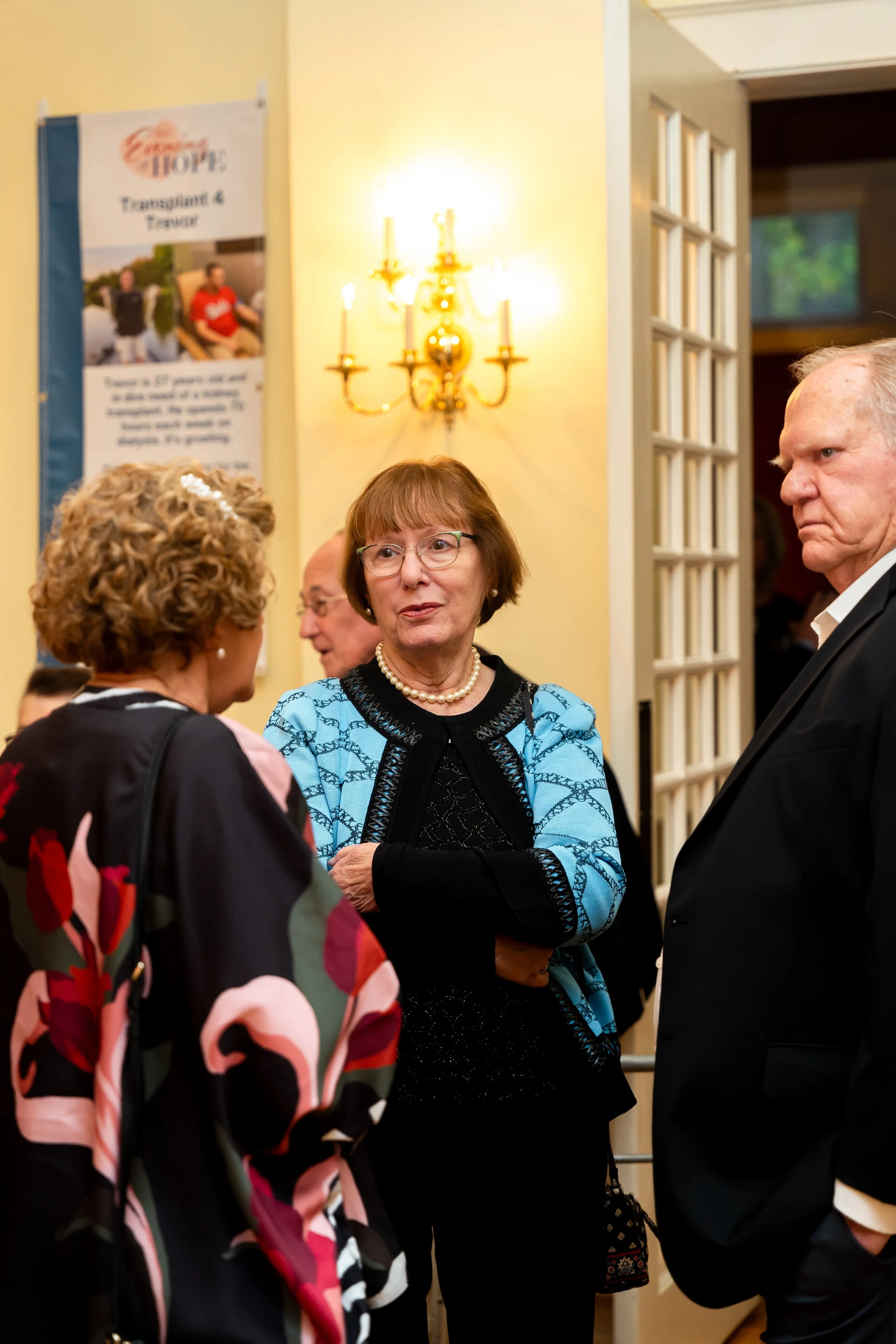 A group of four people having a conversation in a room with yellow walls, with a wall-mounted chandelier, and a poster of a transplant and donor event in the background.