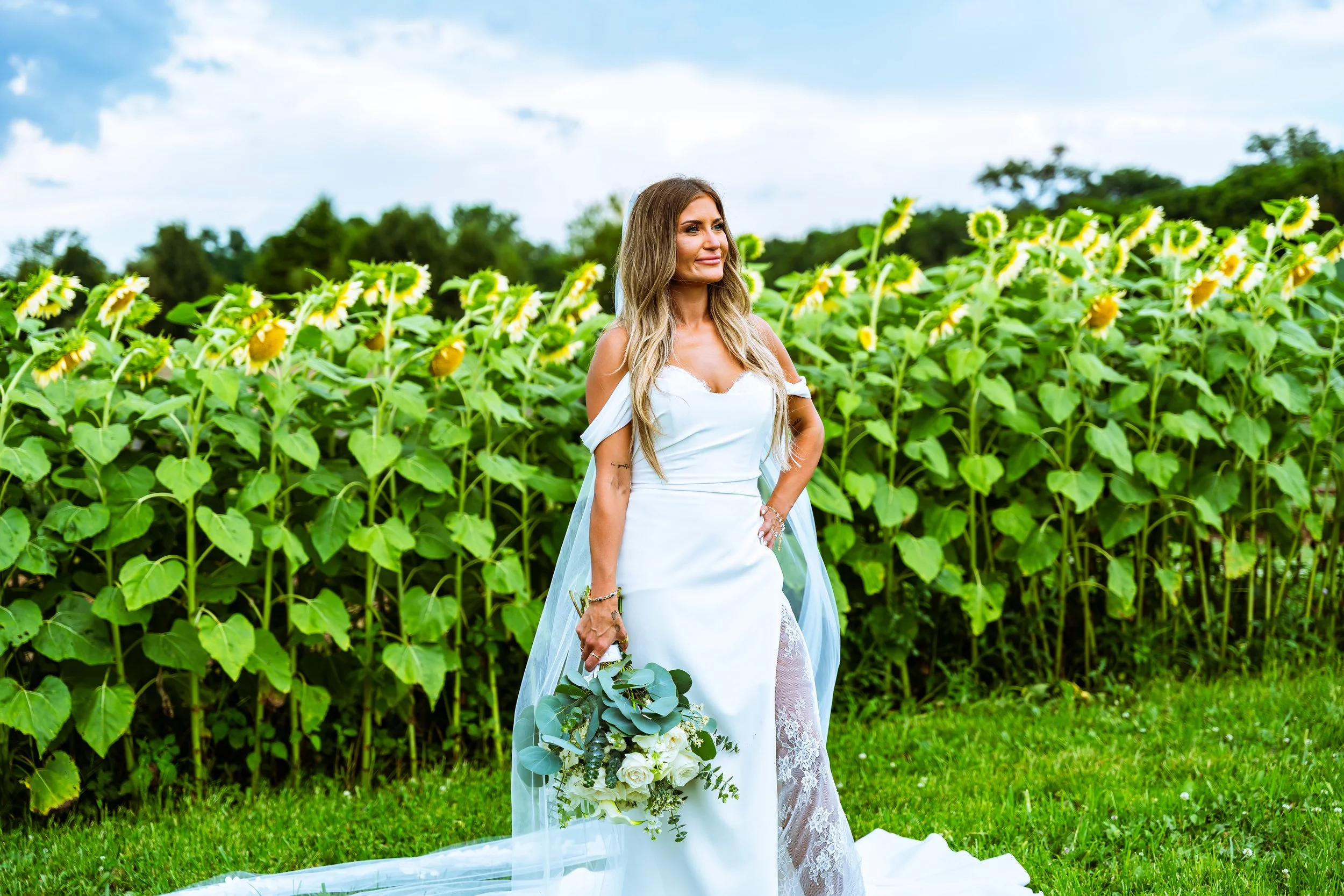 A woman in a white wedding dress holding a bouquet of white roses and greenery, standing in front of a field of sunflowers with a partly cloudy sky overhead.