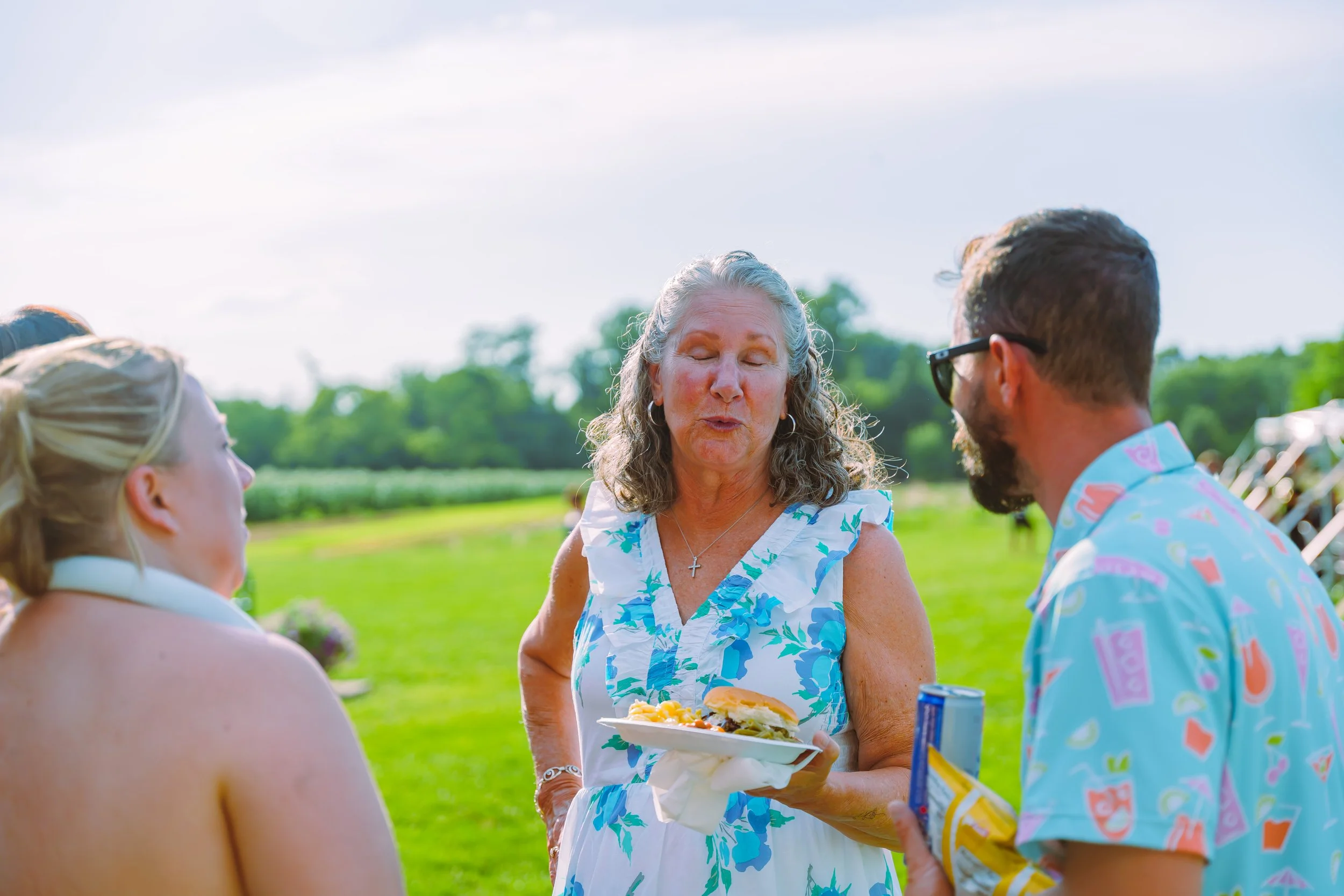 An older woman with gray hair and a blue floral dress is holding a plate of food while talking to three younger people outdoors in a green field under a bright sky.