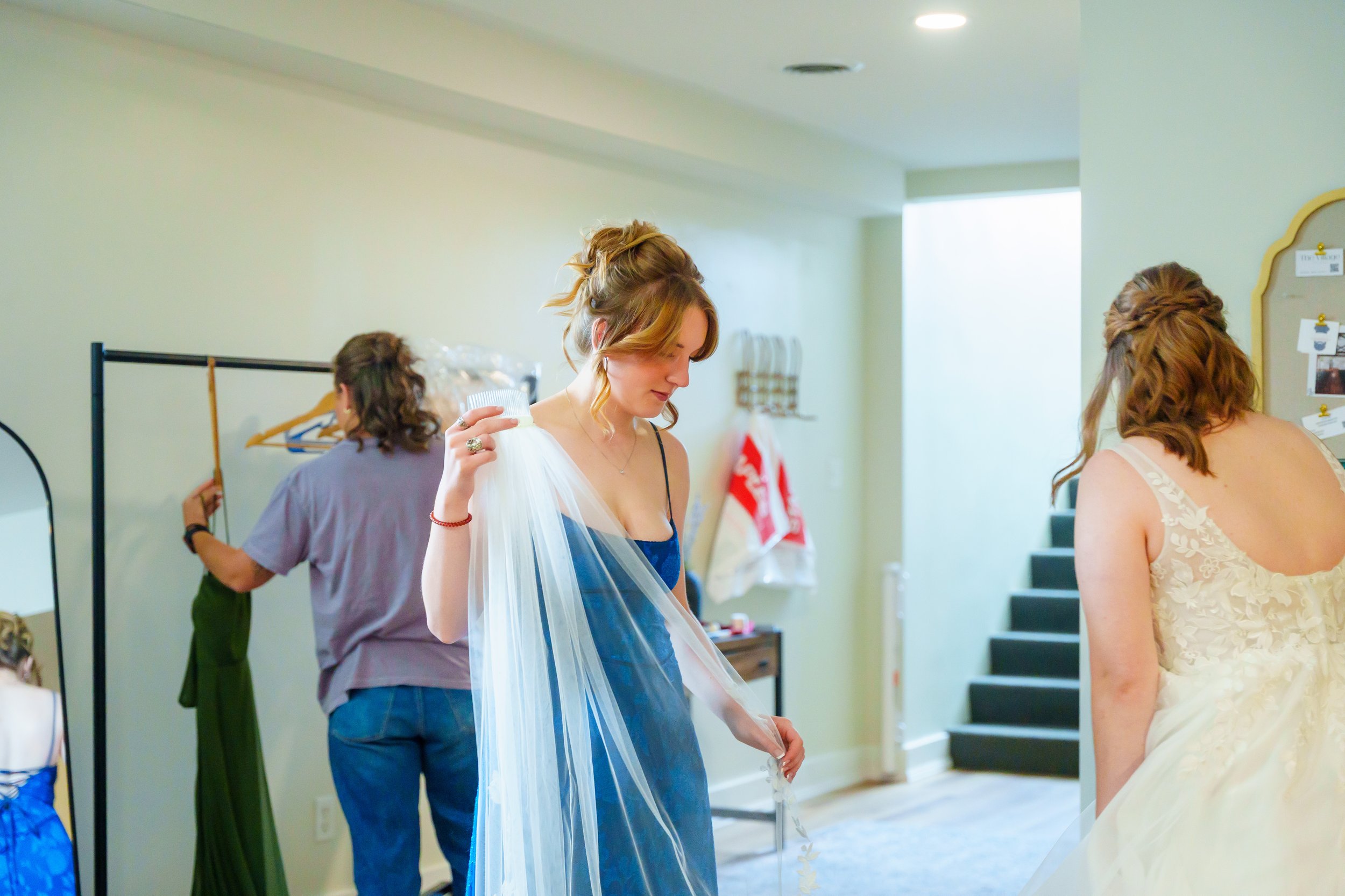 A bride getting ready, holding a piece of sheer fabric, in a room with women preparing behind her, one in a gray shirt and green apron, and two others with their backs turned, wearing dresses, with a staircase and wall decorations visible in the back