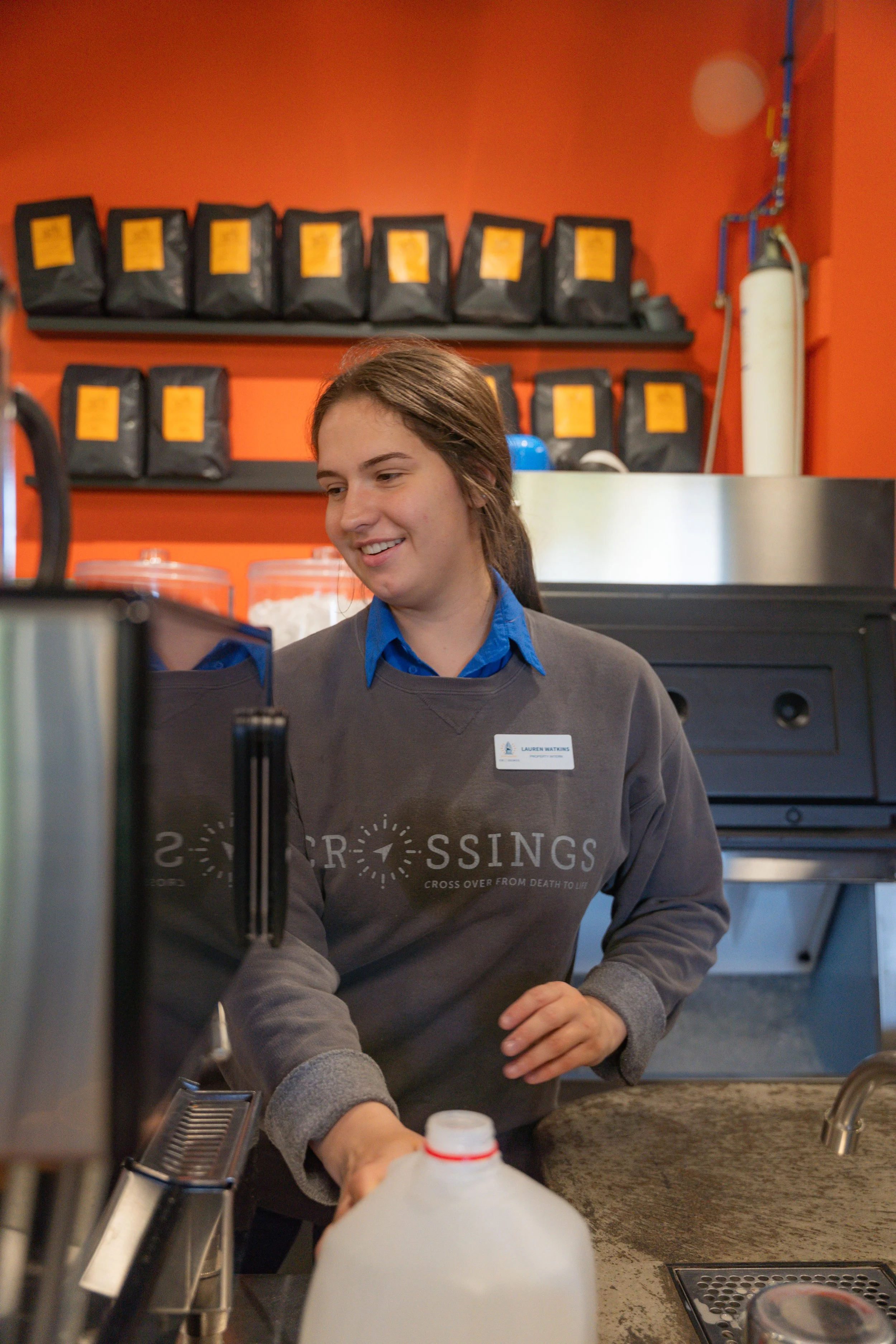 A young woman with long brown hair, wearing a gray sweatshirt and blue shirt, preparing coffee at a coffee shop counter with a cheerful expression, orange wall in the background with black bags of coffee beans on shelves.