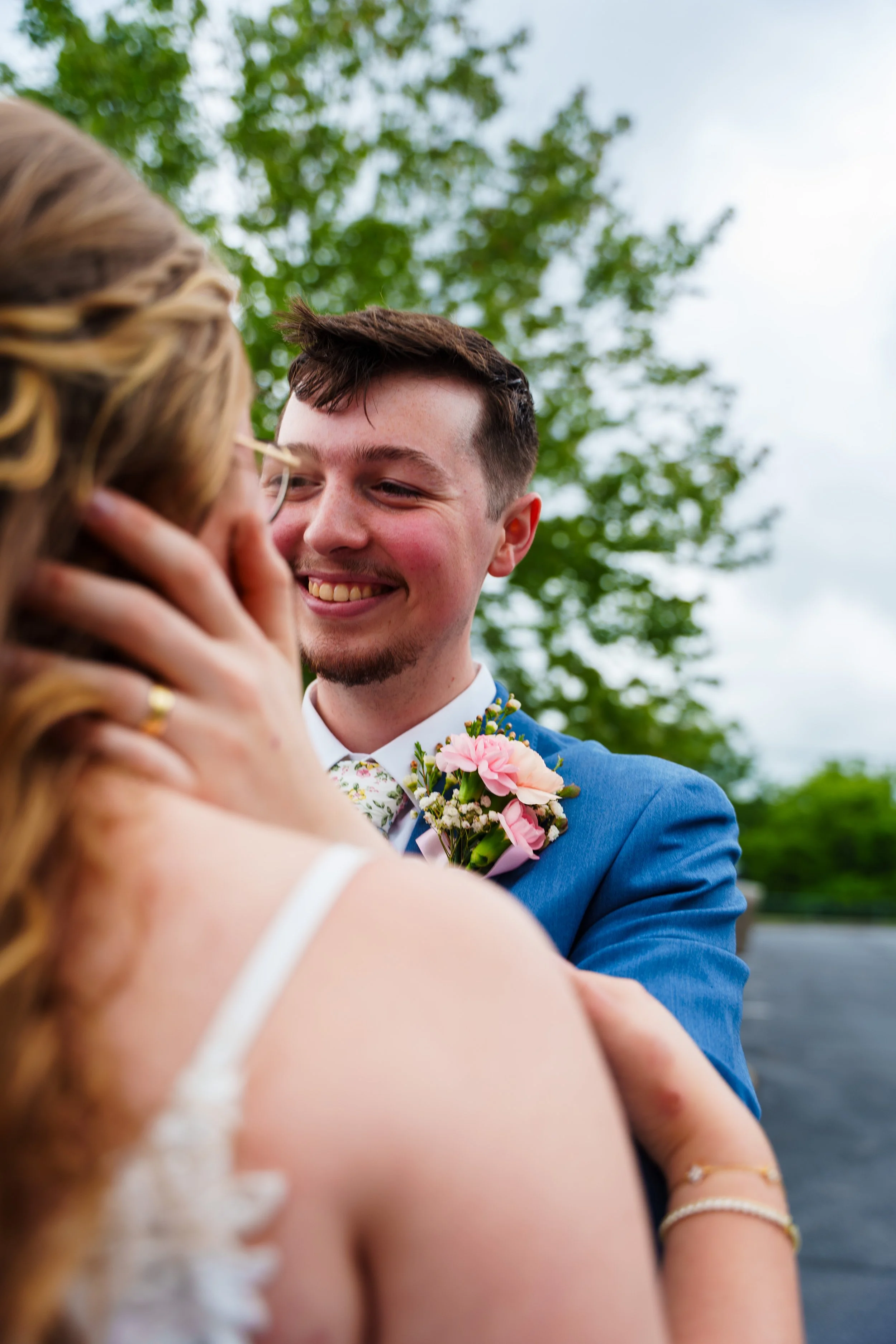 A happy couple, dressed in formal attire, sharing a close moment outdoors with trees and a cloudy sky in the background.