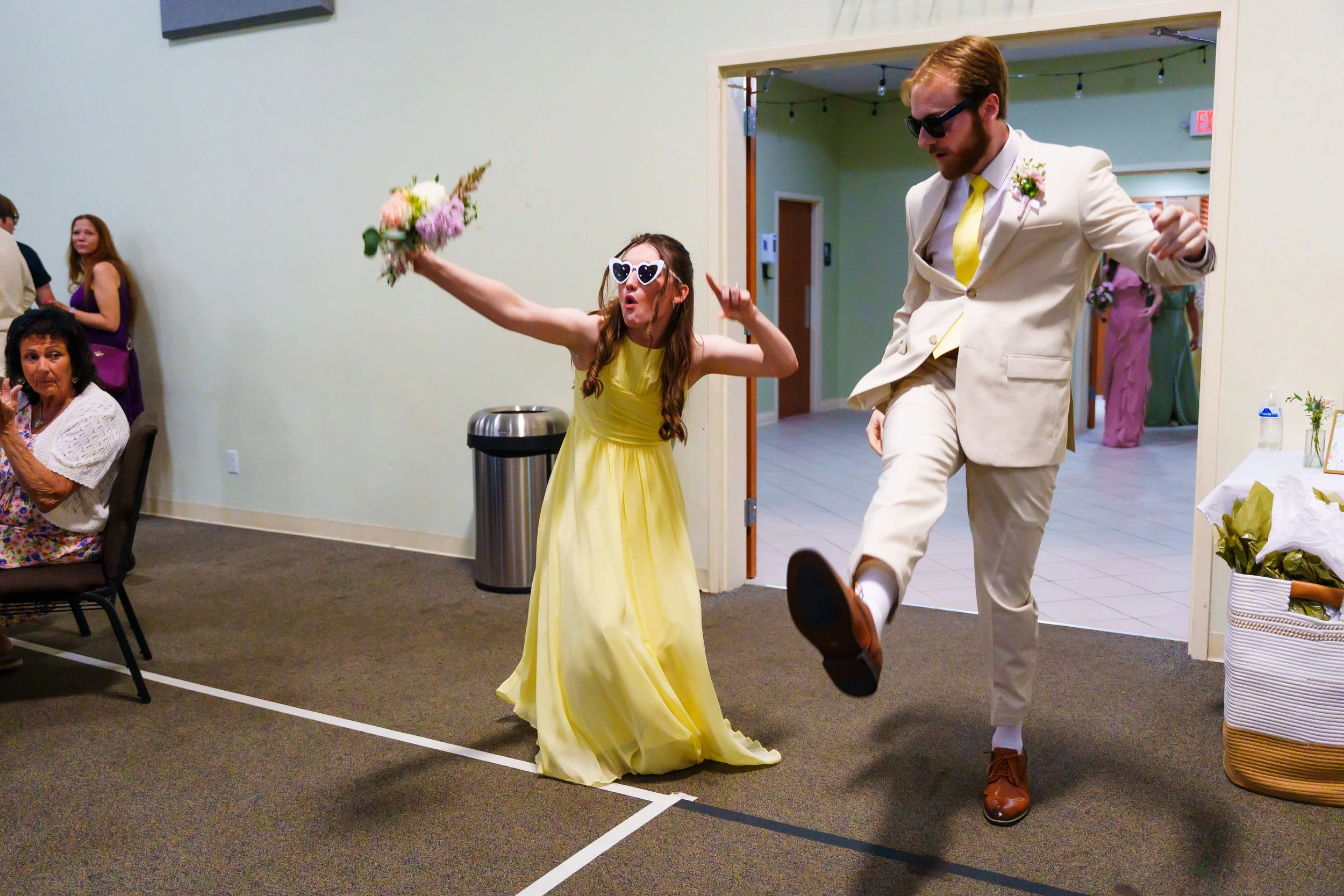 A woman in a yellow dress and heart-shaped sunglasses dancing with a man in a cream suit and black sunglasses at a social event. Other guests are seated and standing around, and there are floral arrangements and a trash bin in the background.