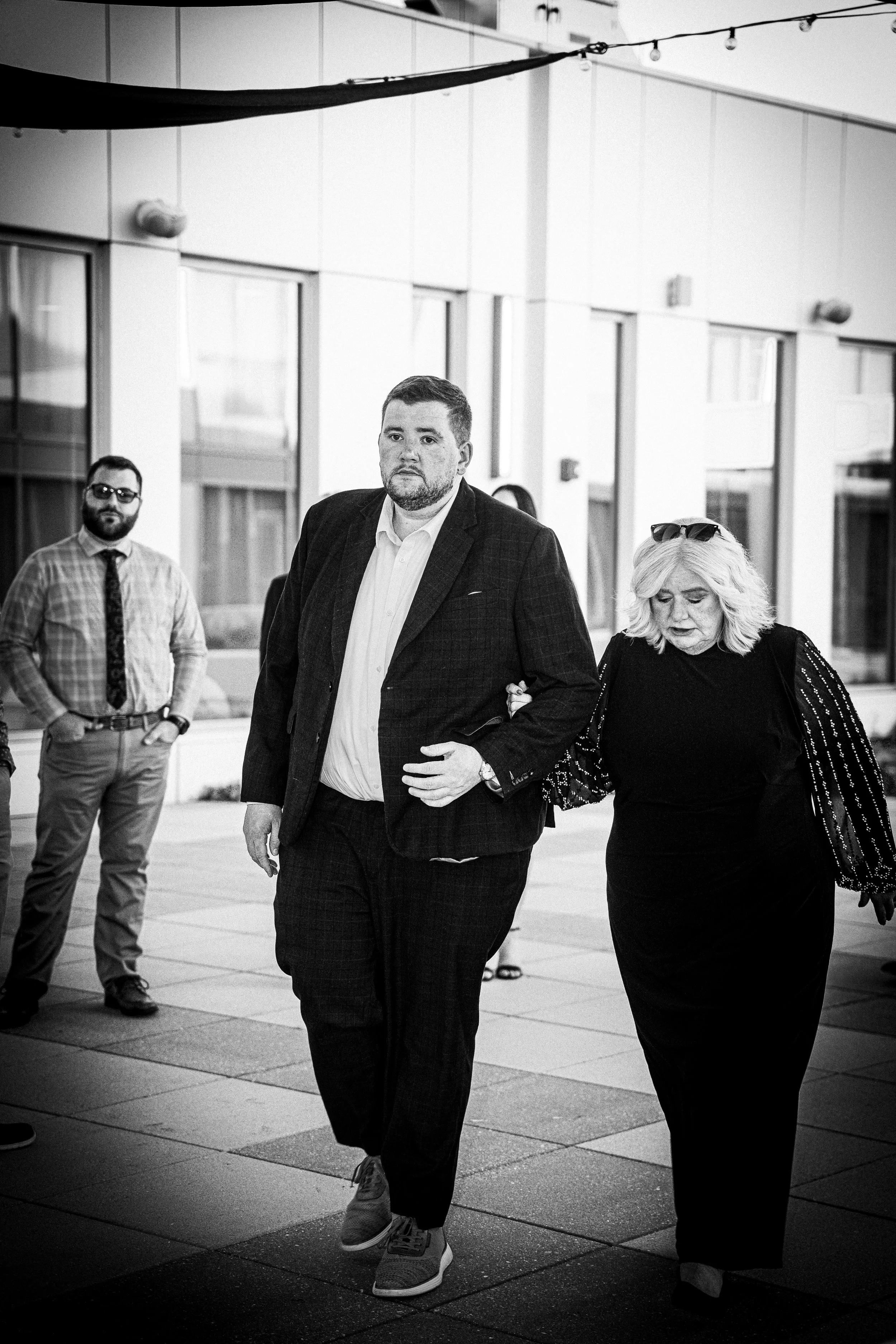 Black and white photo of a man and woman walking outside on a paved area, with a building in the background. The man is in a suit, and the woman is dressed in black with sunglasses on her head. There are other people in the background, and string lig