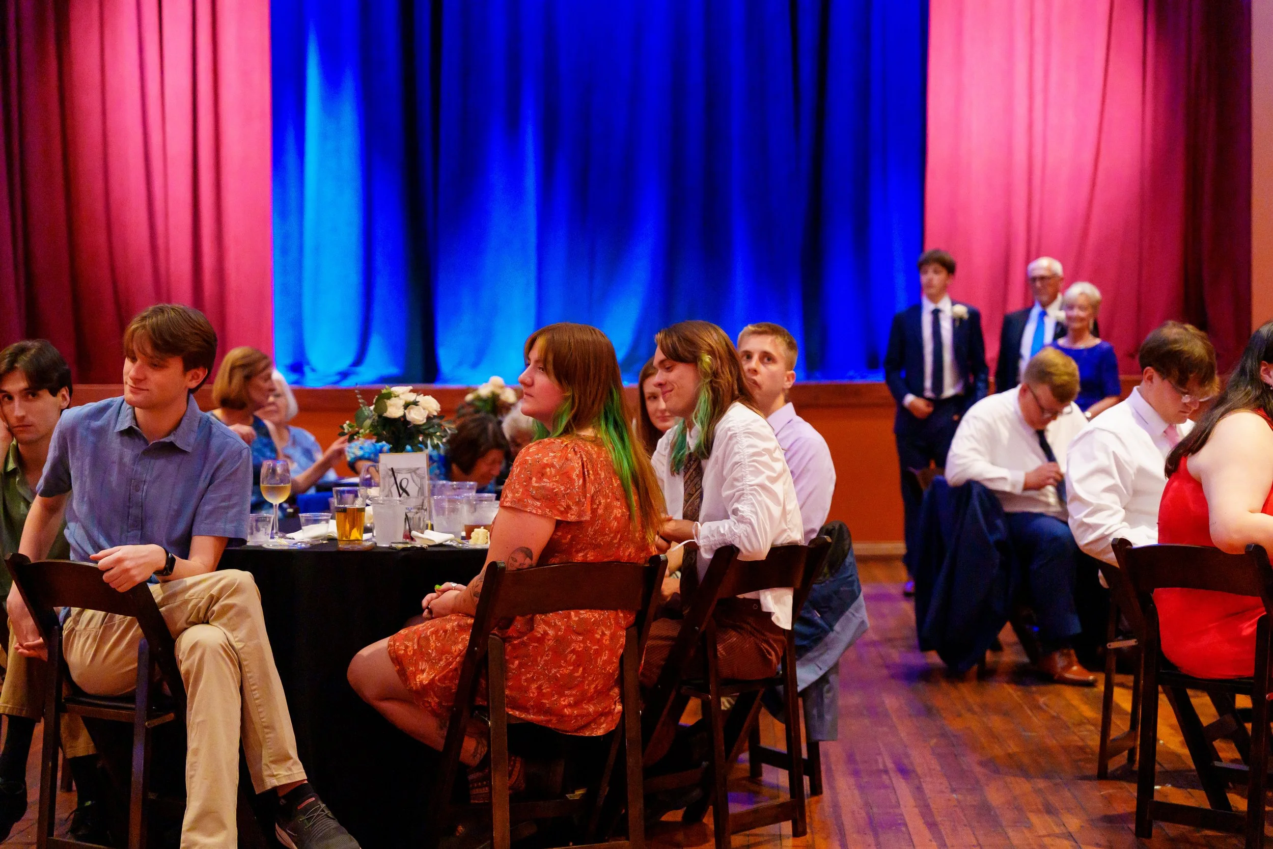 People attending formal event or banquet, seated at round tables with floral centerpieces, in front of stage with blue curtain, some engaged in conversation, others looking around.