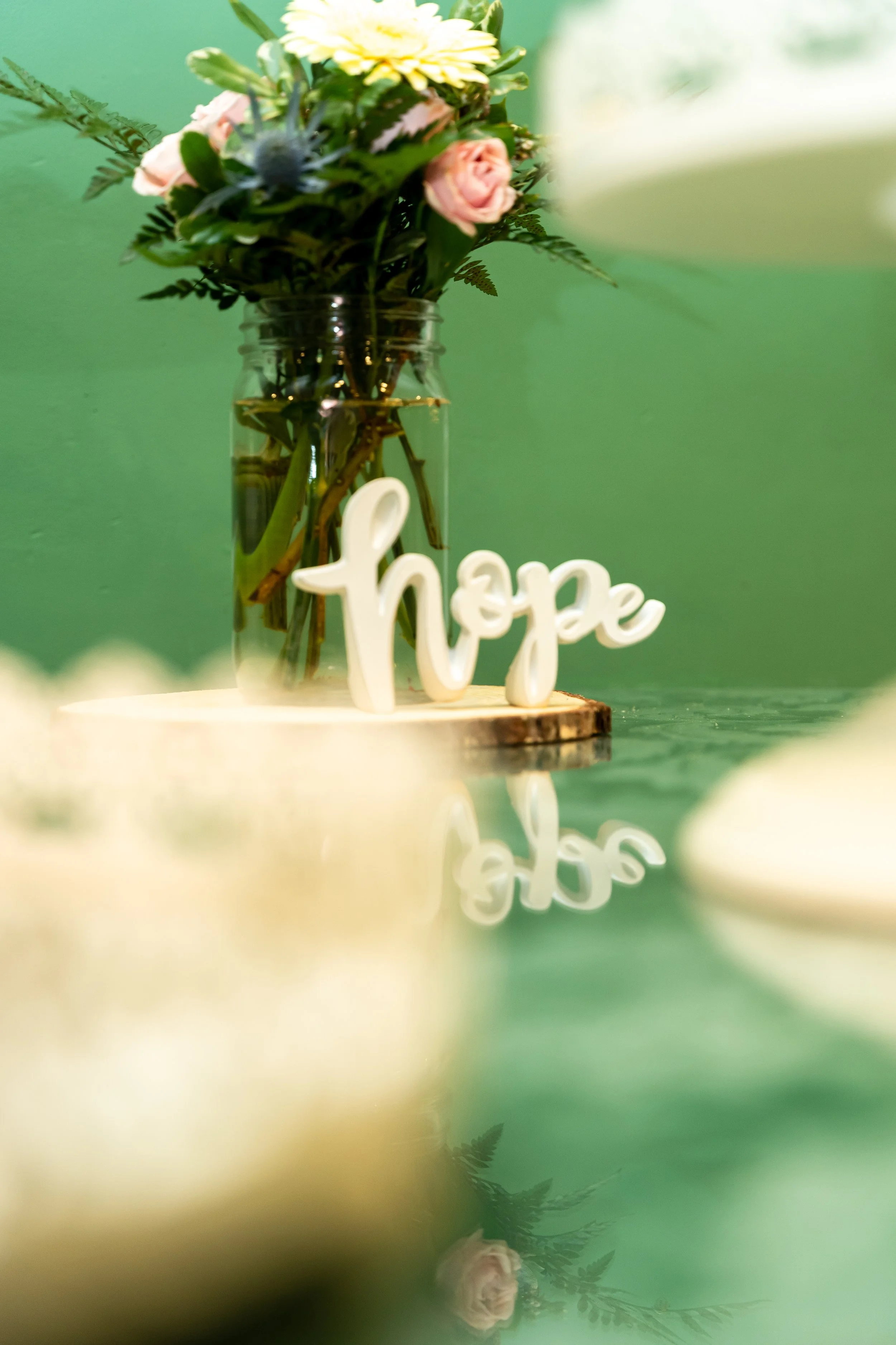 A bouquet of flowers in a glass jar with a white decorative word 'hope' in front, set on a wooden surface with a blurred green background.
