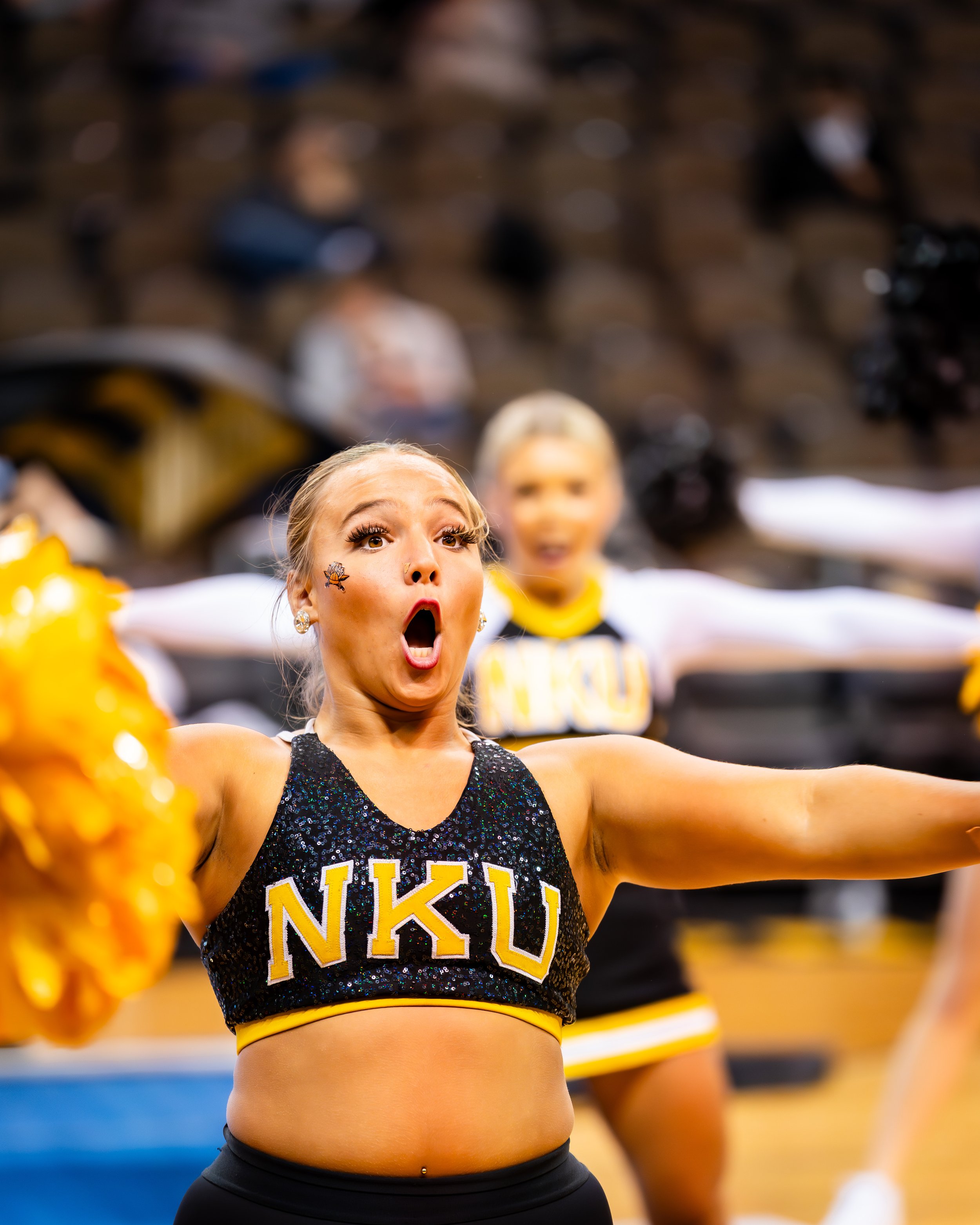 A cheerleader from NKU with a surprised expression, performing at an indoor sports event, with other cheerleaders in the background.