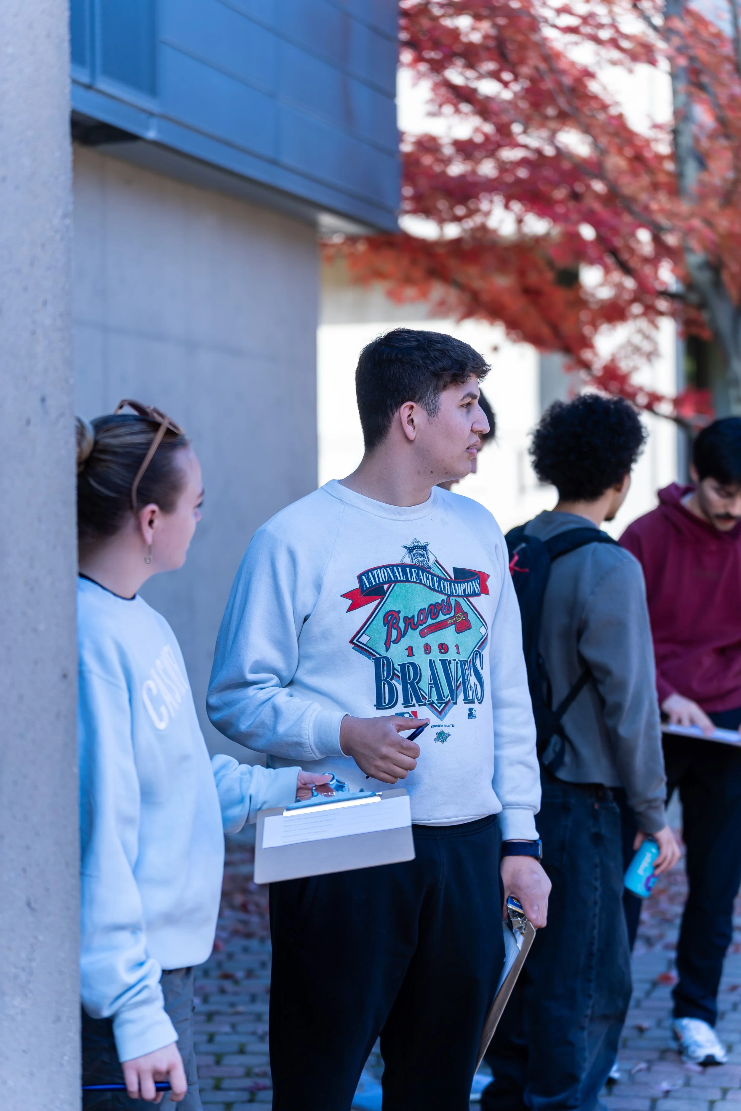 A group of people standing outdoors against a building wall with red autumn leaves on trees in the background.