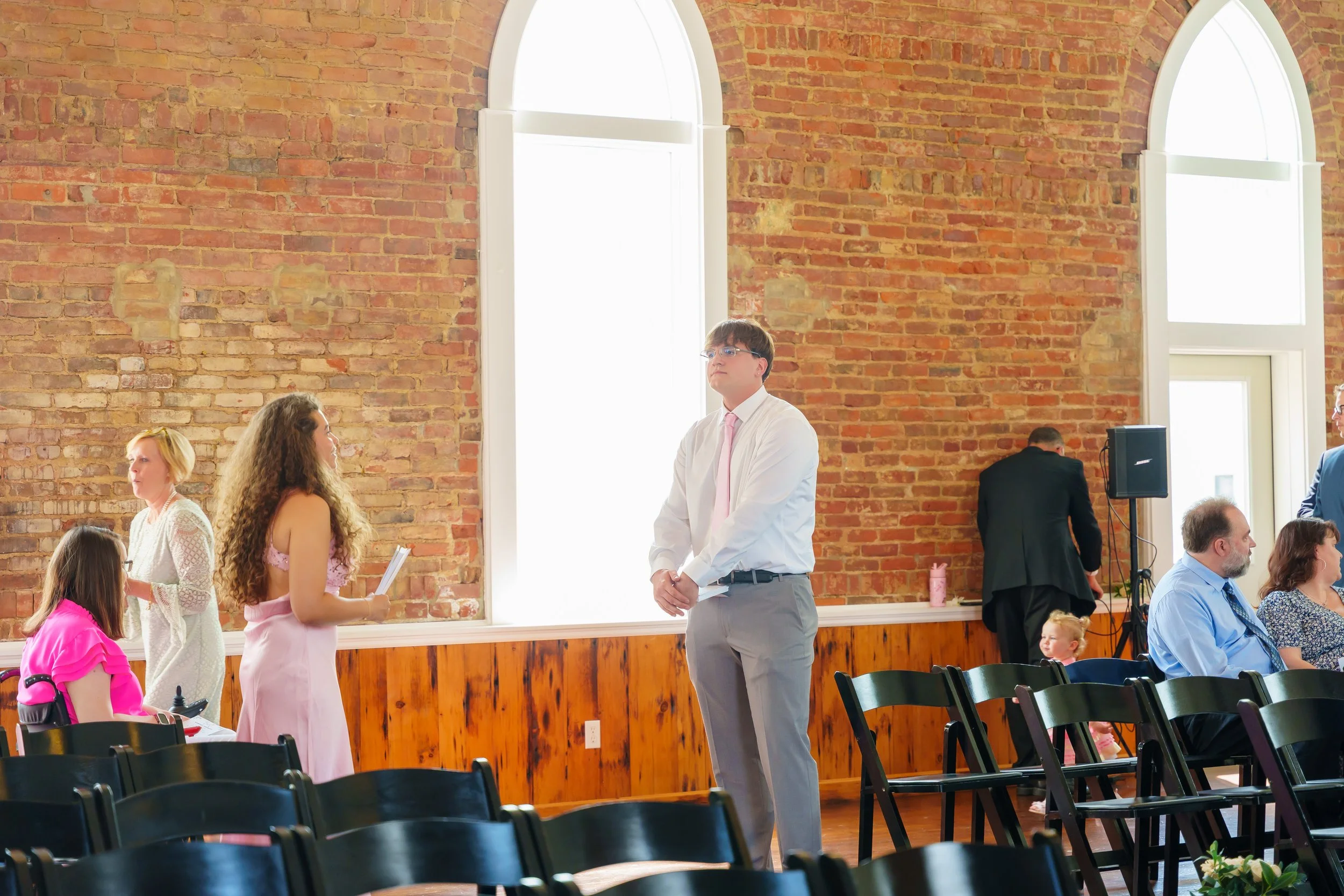 People gathered in an indoor venue with brick walls and tall arched windows, engaged in conversation and possibly waiting for a presentation or event to start.
