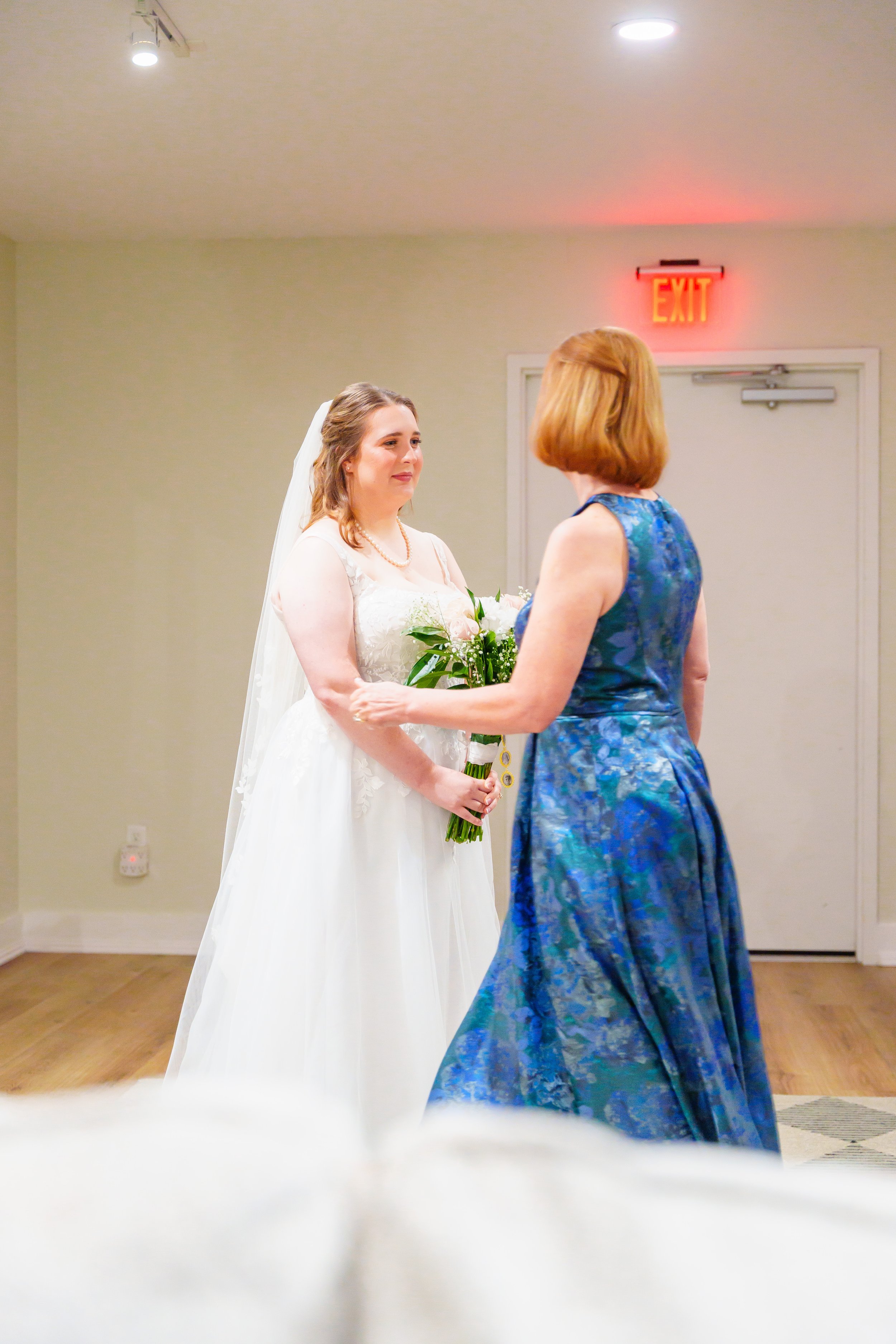 A bride and an older woman are facing each other indoors in a wedding setting. The bride holds a bouquet of flowers and is listening to the woman. An exit sign is above a door in the background.