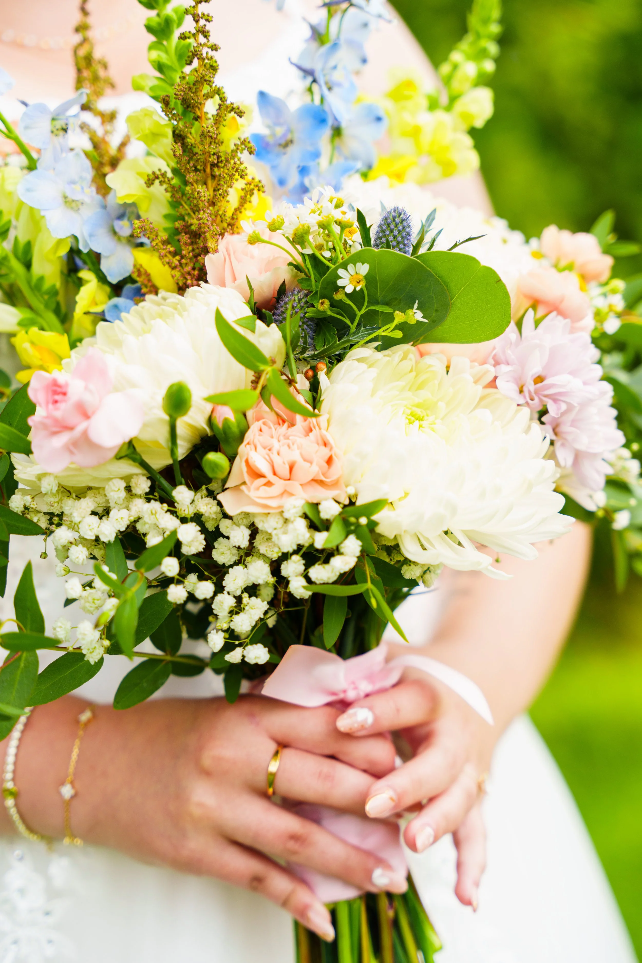 A person holding a colorful bouquet of flowers including white, pink, blue, and yellow blooms, with green leaves, and a pink ribbon around the stems.
