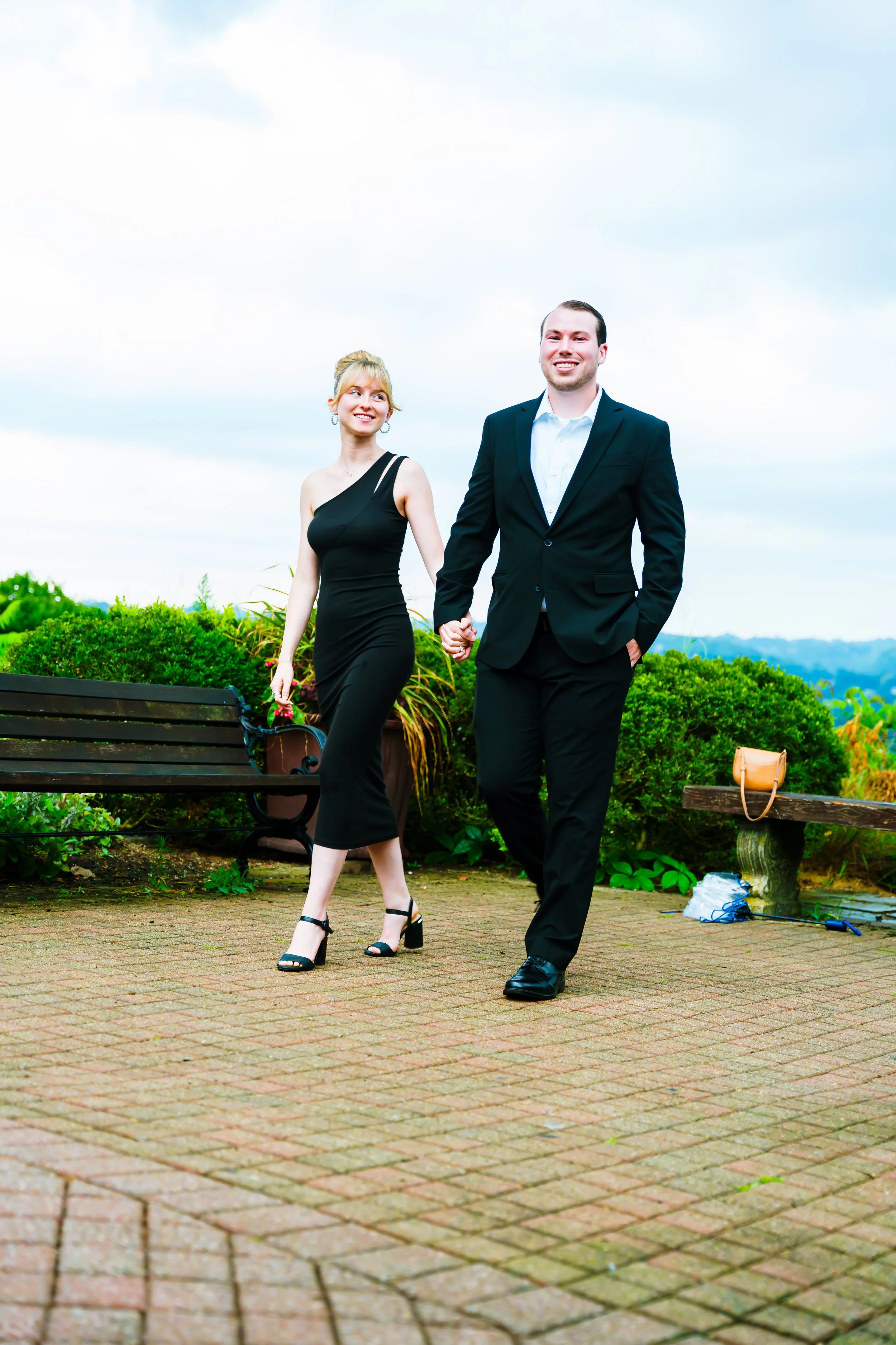 A couple holding hands walking in a park with bench, greenery, and cloudy sky in the background.