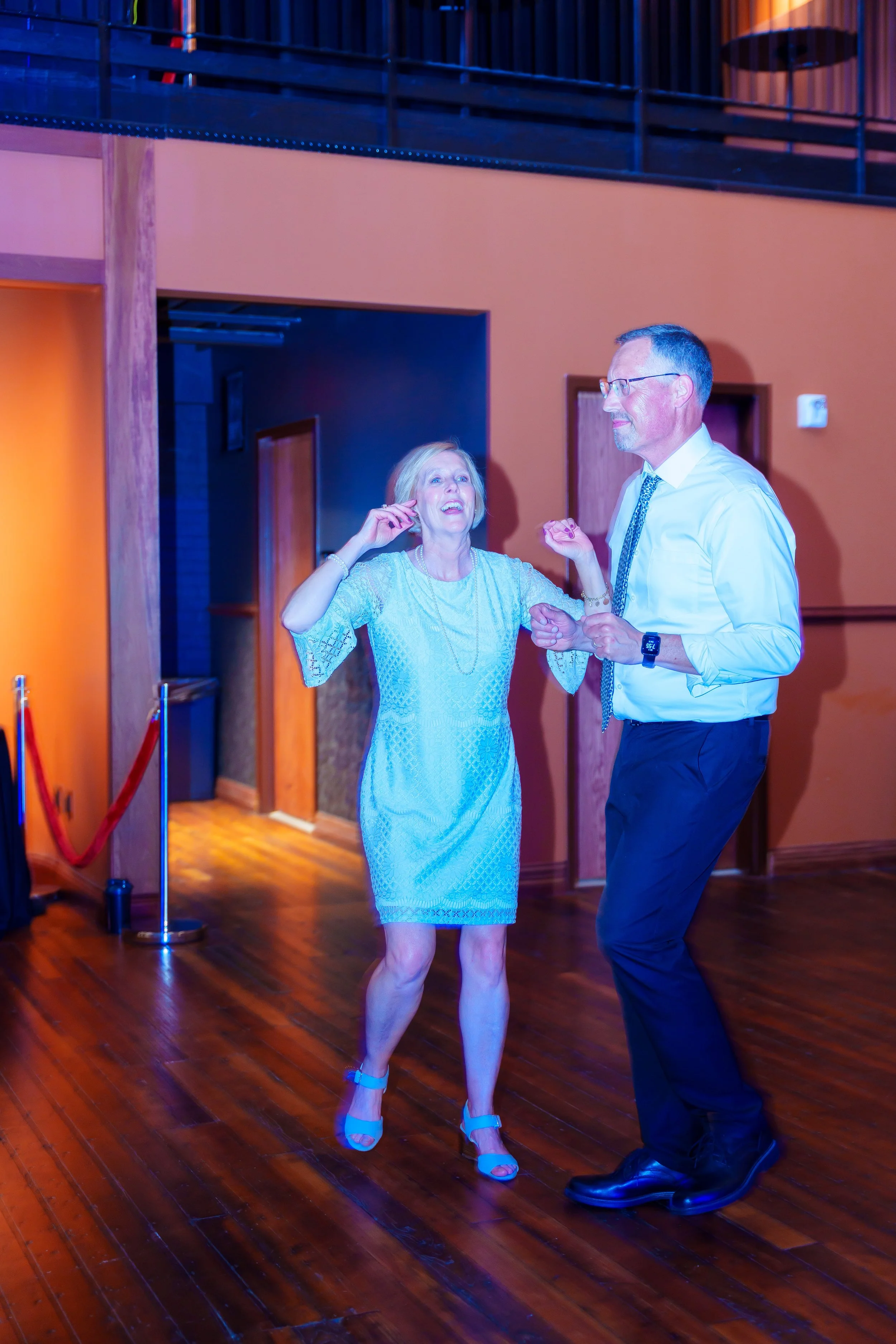 A woman in a light blue lace dress and heels is dancing and smiling with a man in a white shirt and black pants, both enjoying themselves at an indoor event with wooden floors and warm-colored walls.