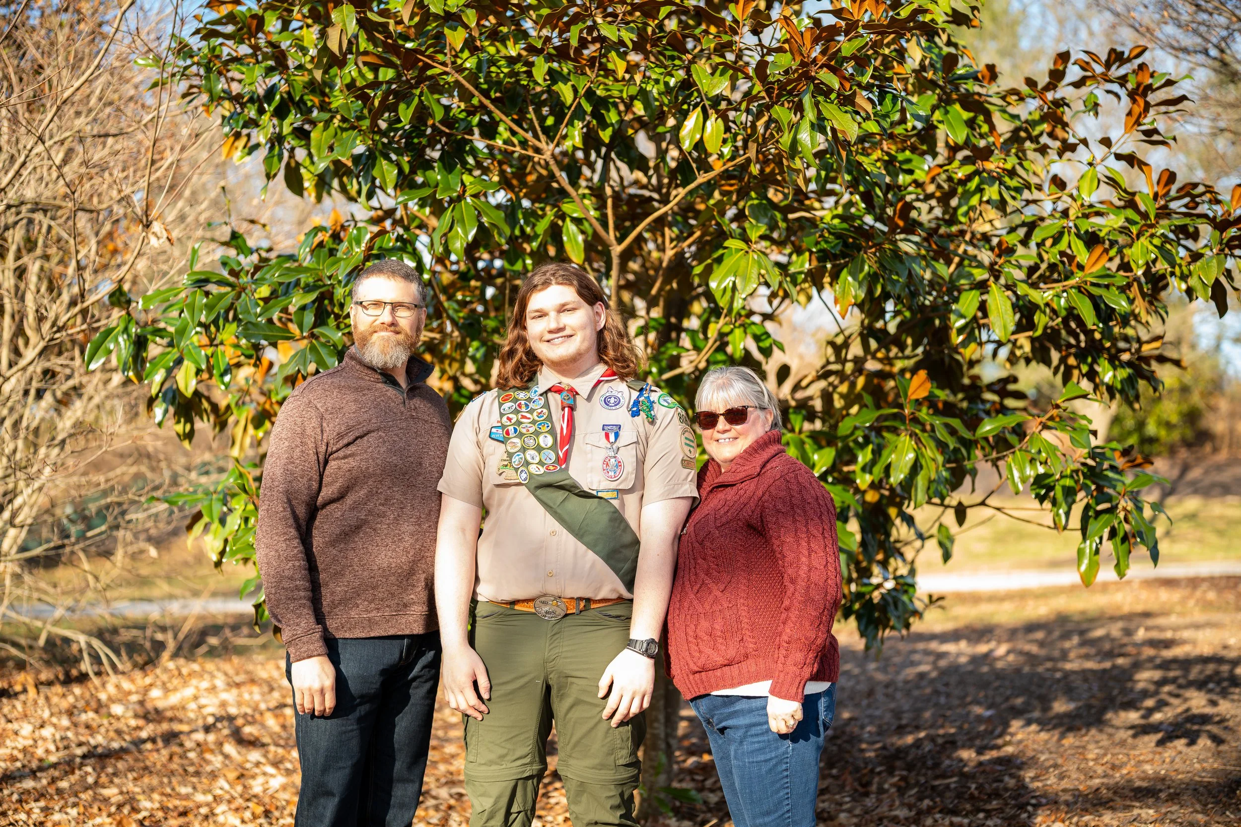 A family of three stands outdoors in front of a large green tree, smiling at the camera. The young man in the middle is a Boy Scout in uniform with many badges. The woman on the right is wearing sunglasses and a red sweater, and the man on the left is dressed casually in a brown sweater and jeans.