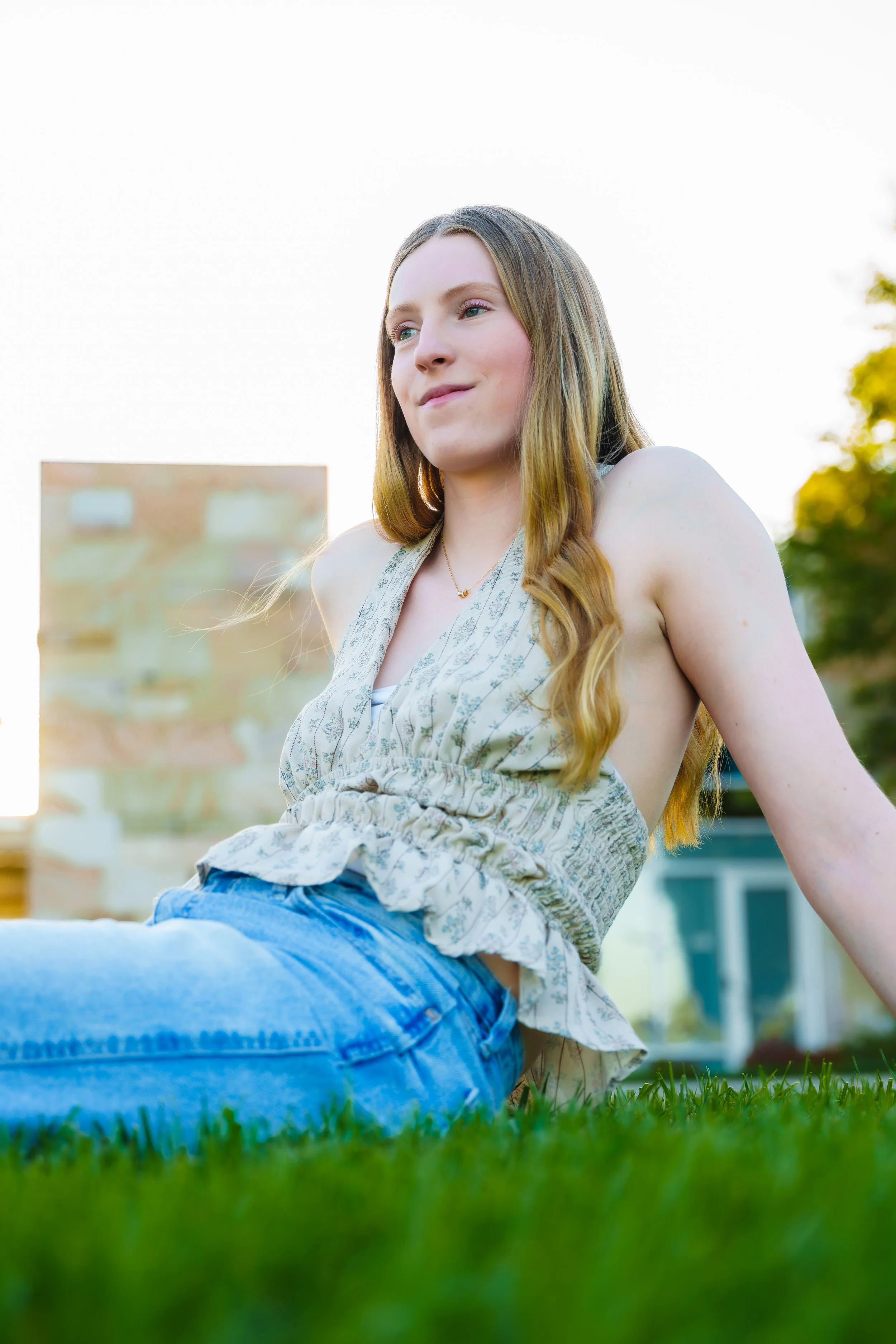 A young woman with long light brown hair, wearing a sleeveless floral top and blue jeans, sitting on the grass outdoors during daylight. She has a slight smile and is looking off into the distance with a calm expression.