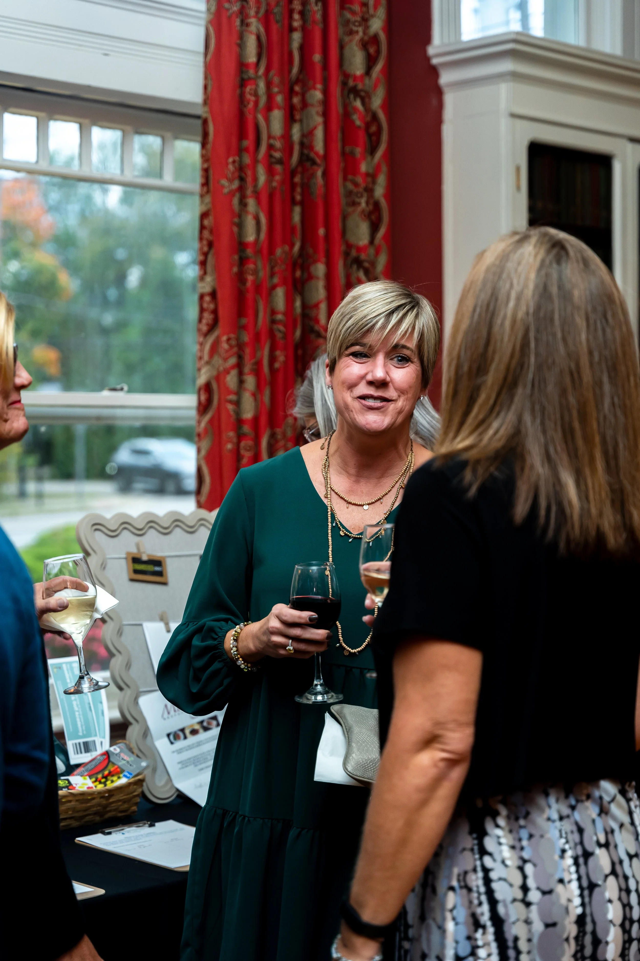 Women talking and holding glasses of wine at a social event indoors near a window with red curtains.