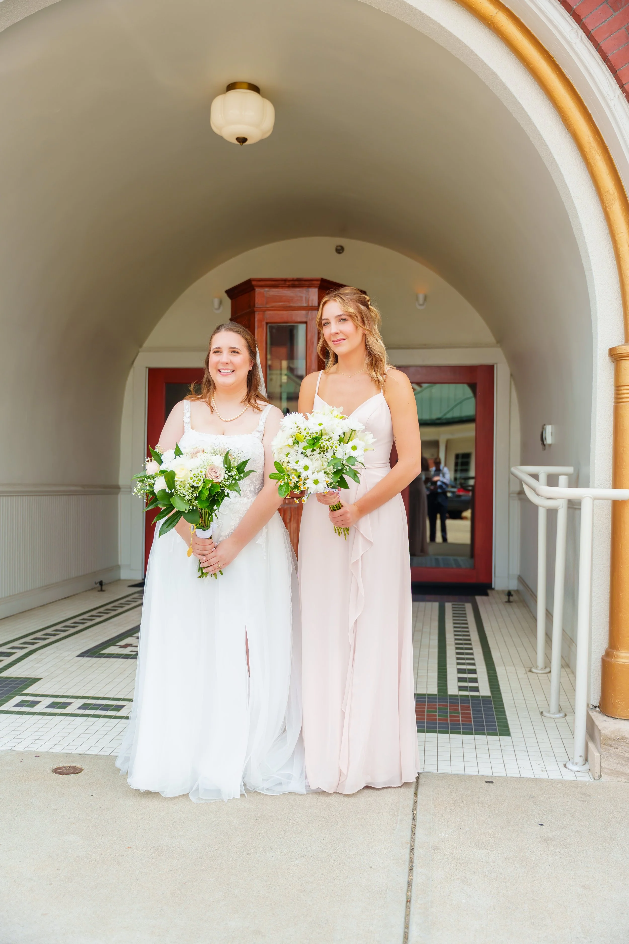 Two women dressed in white dresses holding bouquets, standing under an arched doorway of a building with vintage interior, smiling at the camera.