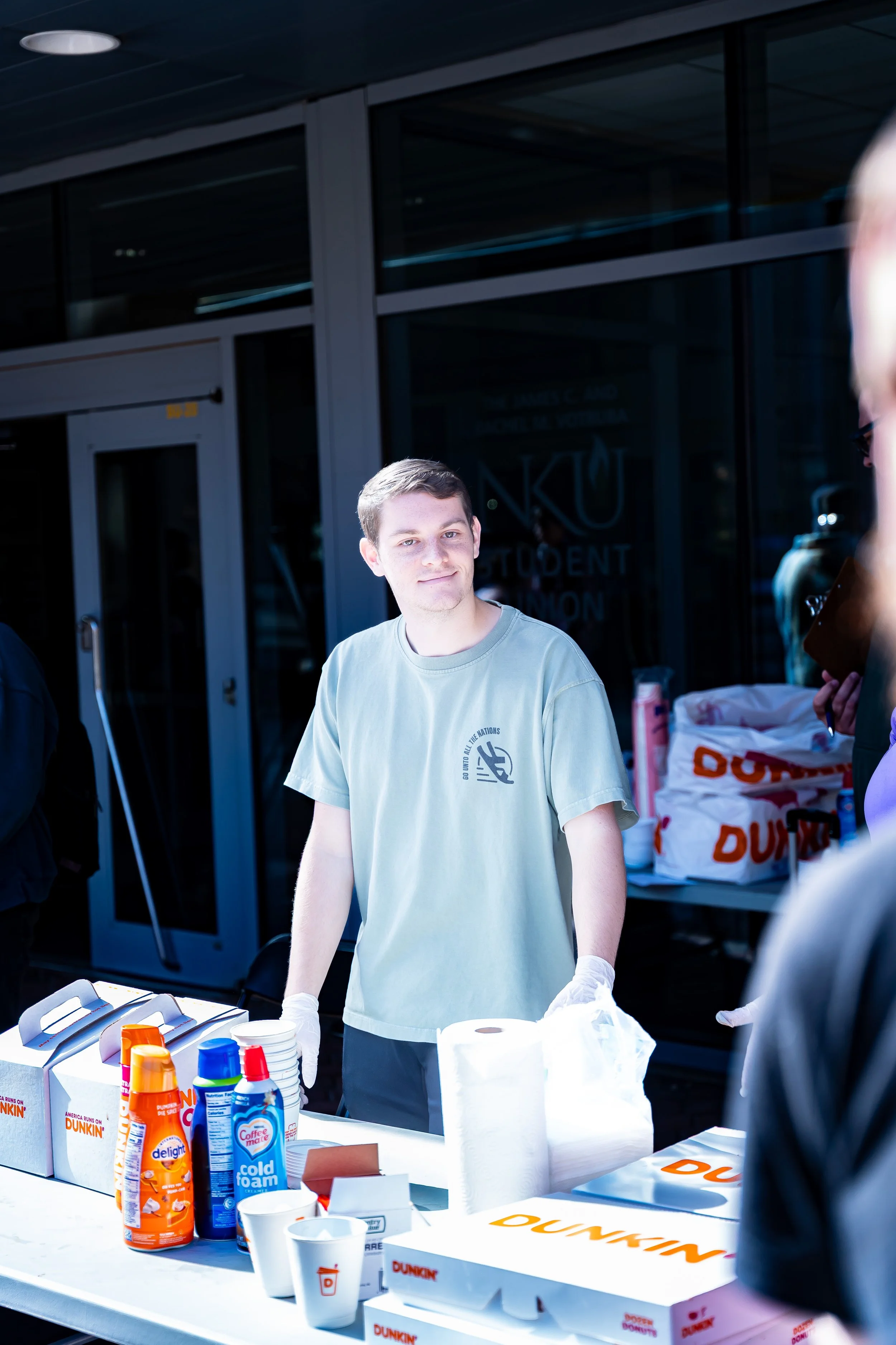 A young man wearing a light gray t-shirt and white gloves standing behind a table with cups, bottles of sauces, and napkins, possibly at a Dunkin' Donuts or Dunkin' event, outdoors near a building entrance.