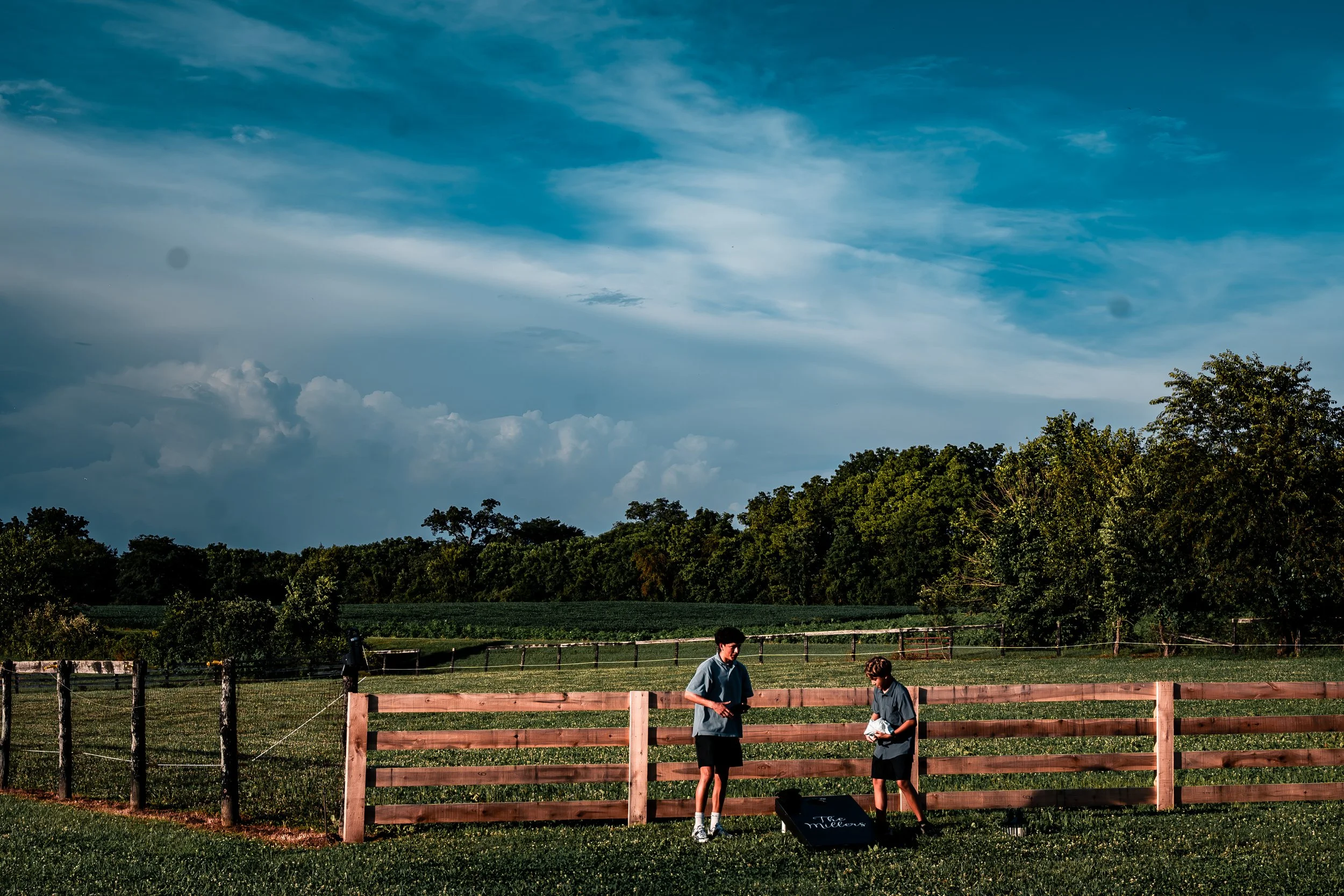 Two boys standing next to a wooden fence in a grassy field with trees, under a partly cloudy sky.