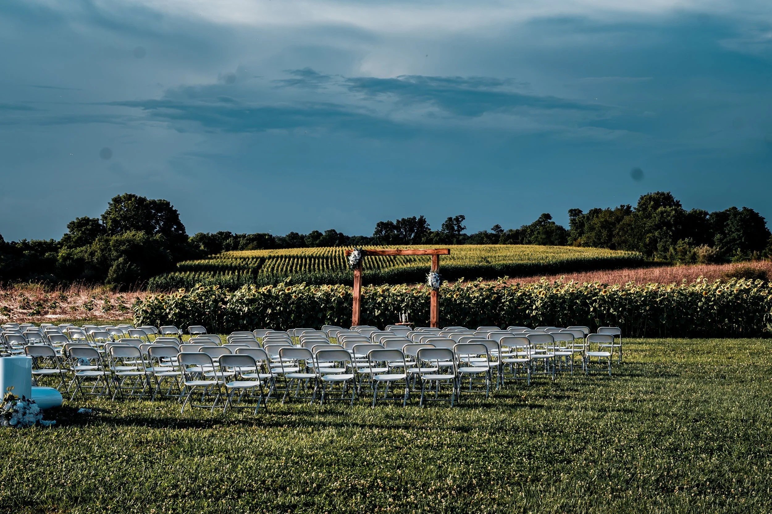 Outdoor wedding ceremony setup with rows of white chairs overlooking a field with crops, under a cloudy sky.