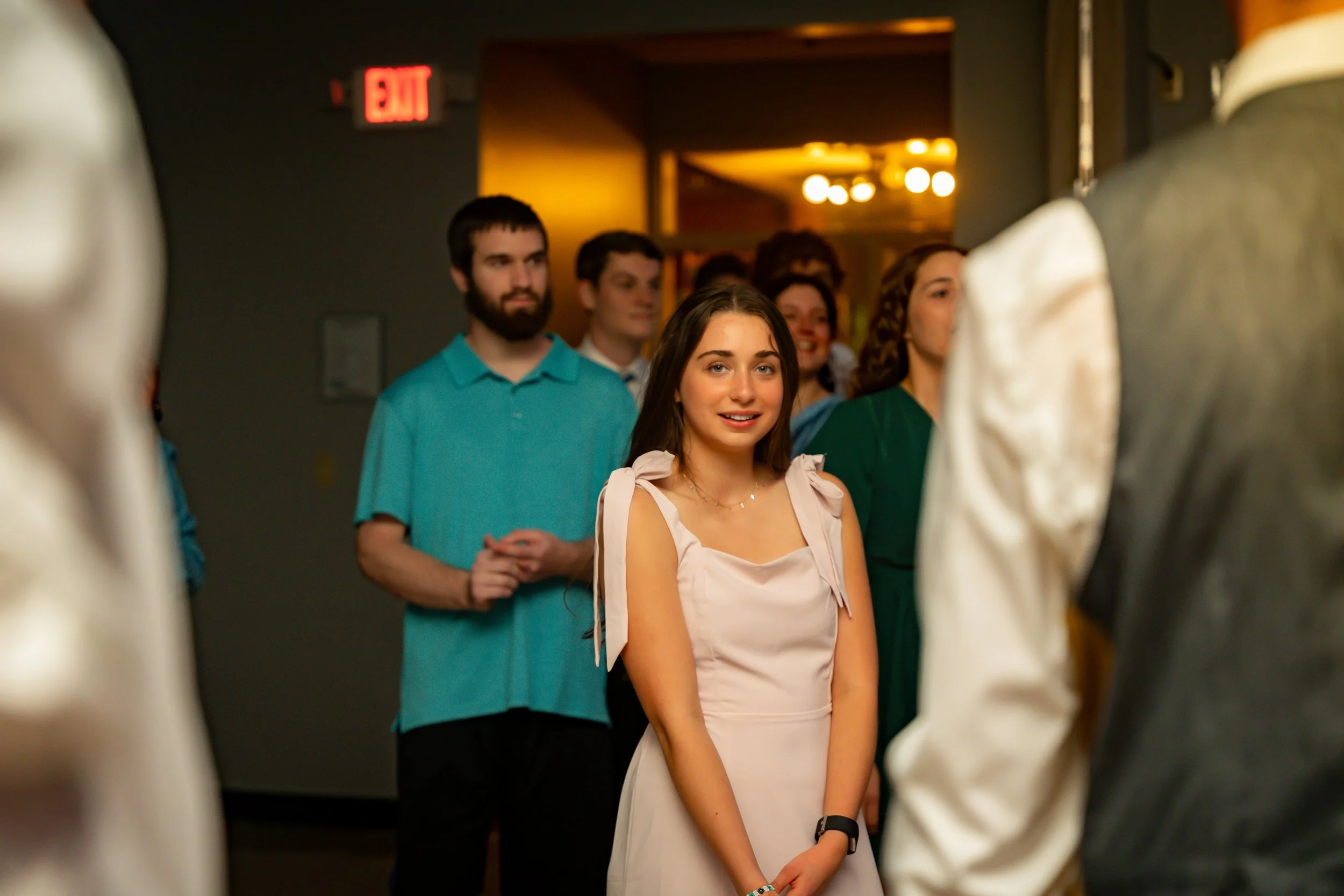 A group of people, mostly young adults, standing in line, with a young woman in front, smiling slightly, wearing a light pink dress with bows on the shoulders, in an indoor setting with warm lighting and an exit sign visible in the background.