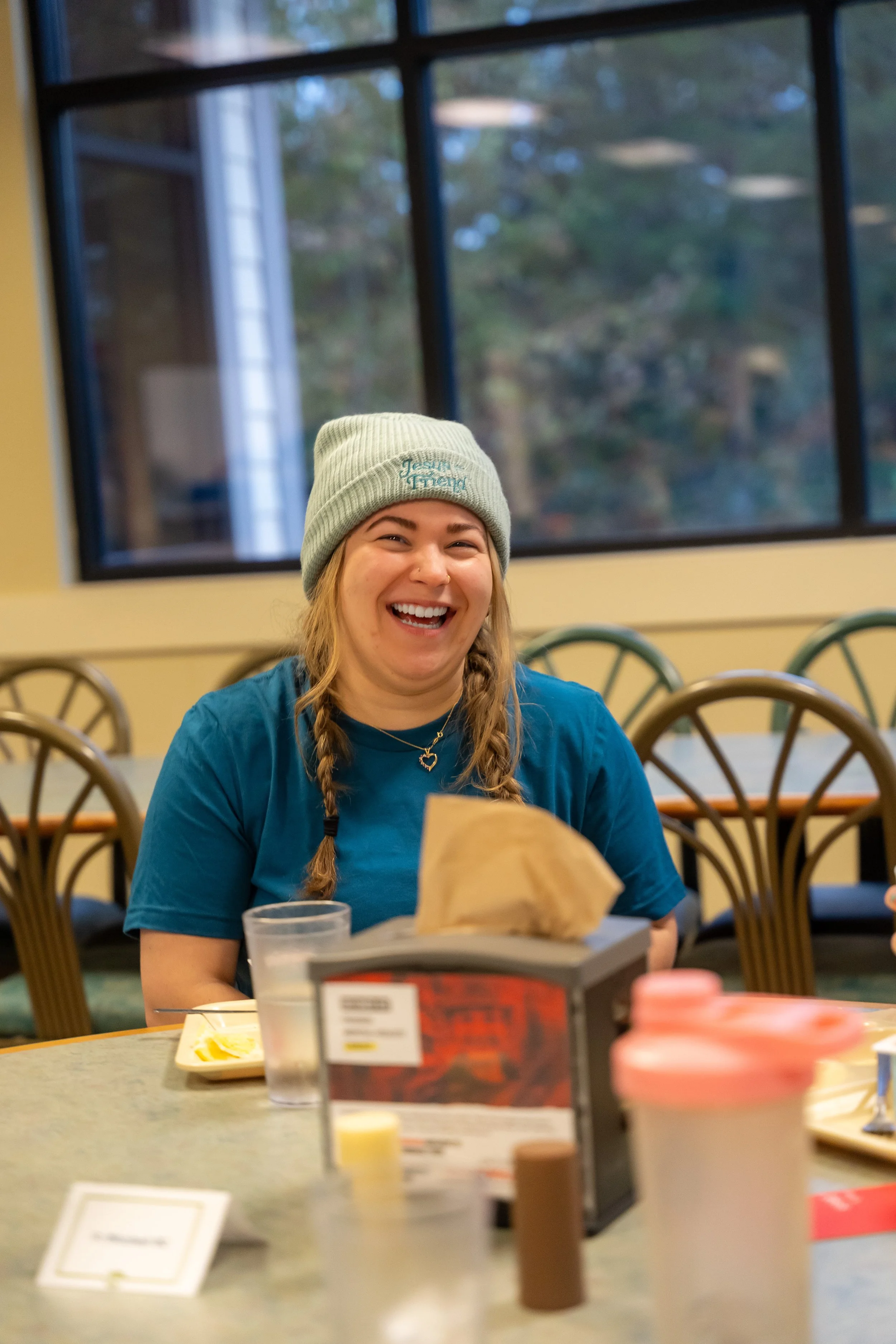 A young woman with braided hair, wearing a gray beanie and blue shirt, smiling at a table in a restaurant or cafe. There are chairs, a menu, condiments, and a glass of water on the table, with large window panes and trees visible outside.