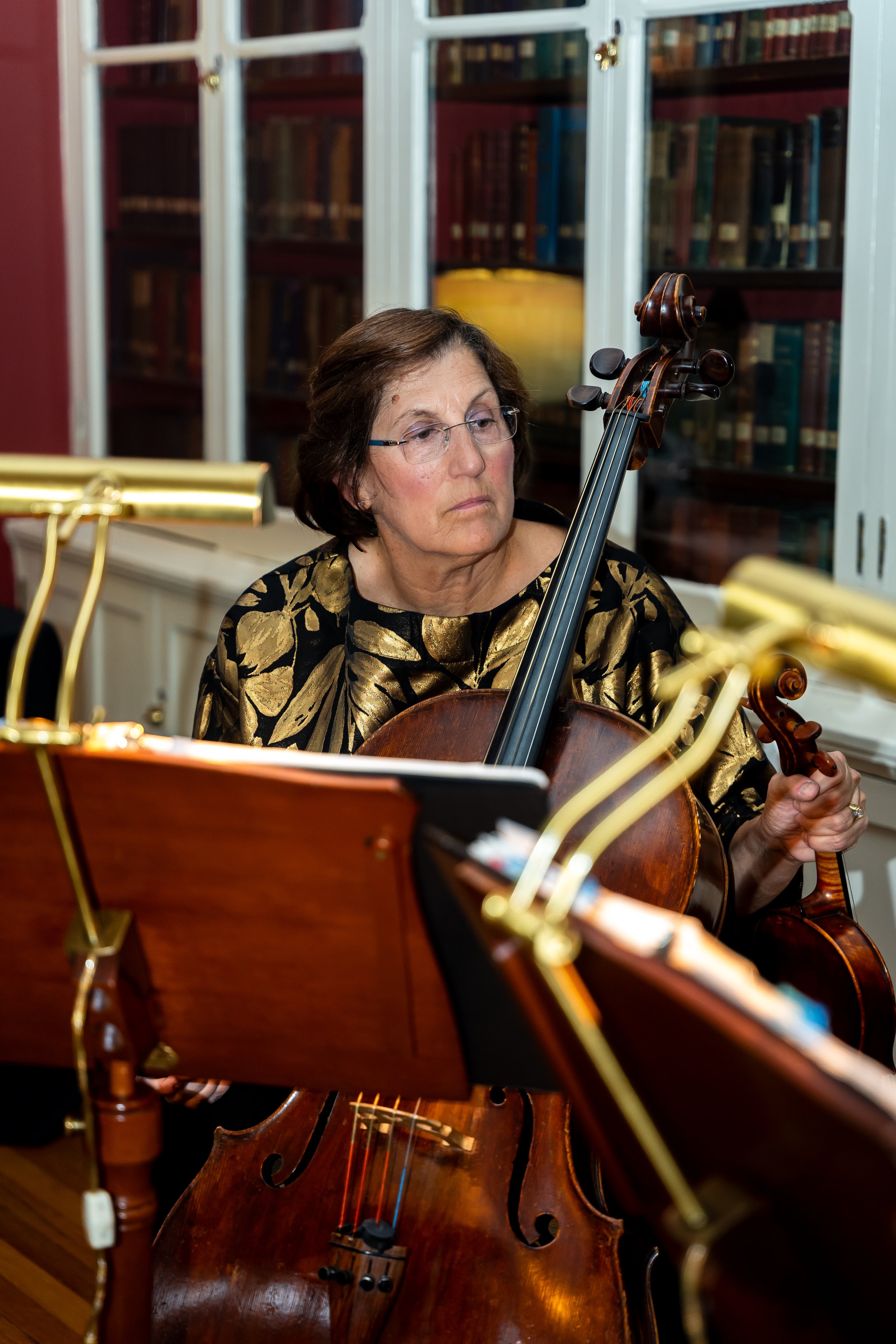 A woman with glasses playing a cello, with sheet music and a music stand in front of her, set against a background of a library with glass-fronted bookshelves.