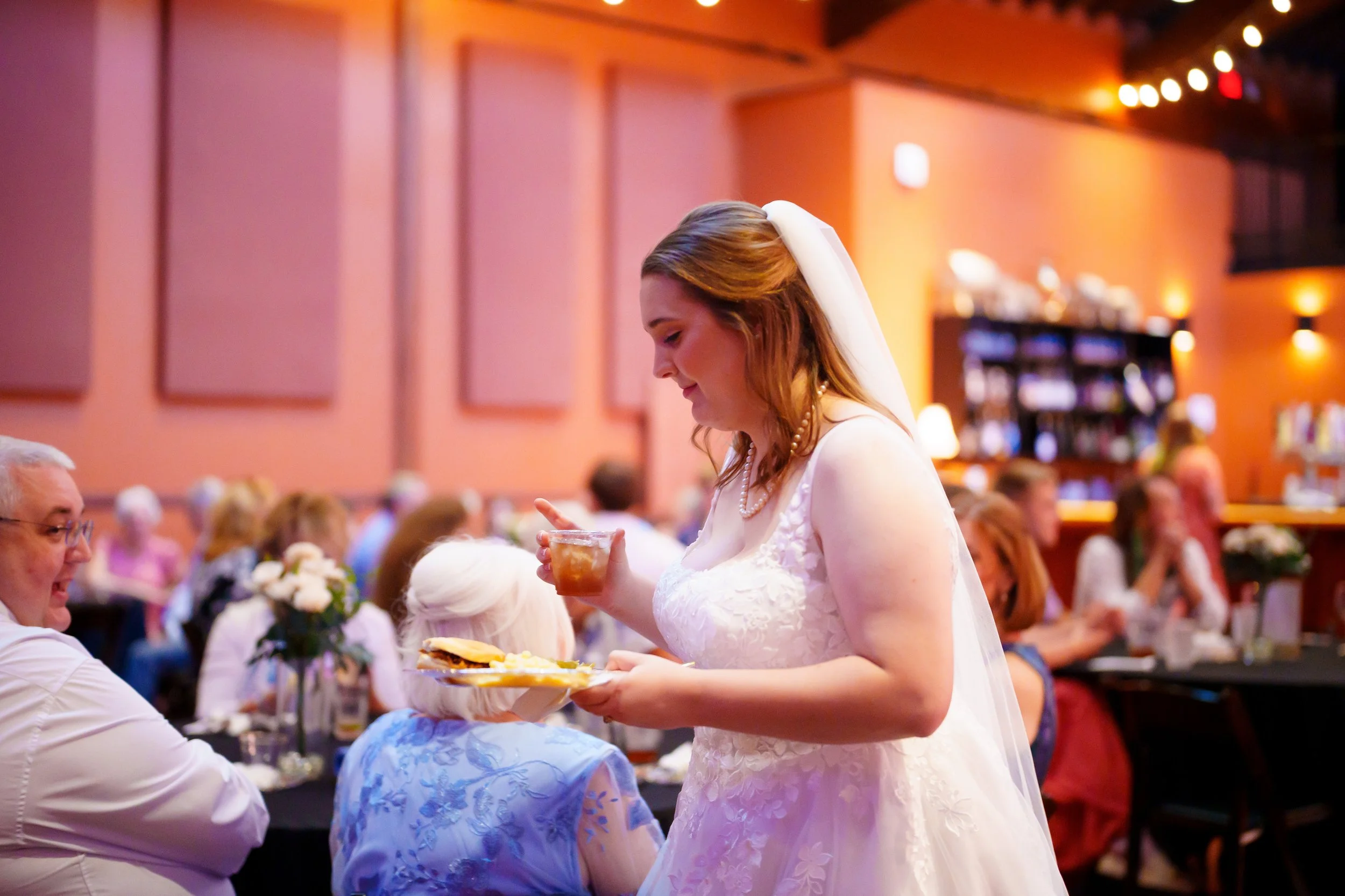 A bride wearing a white wedding dress and pearl necklace is serving food at a reception. She is holding a plate with a sandwich and other items, and a guest is receiving her food. The reception hall is decorated with warm lighting and floral centerpi