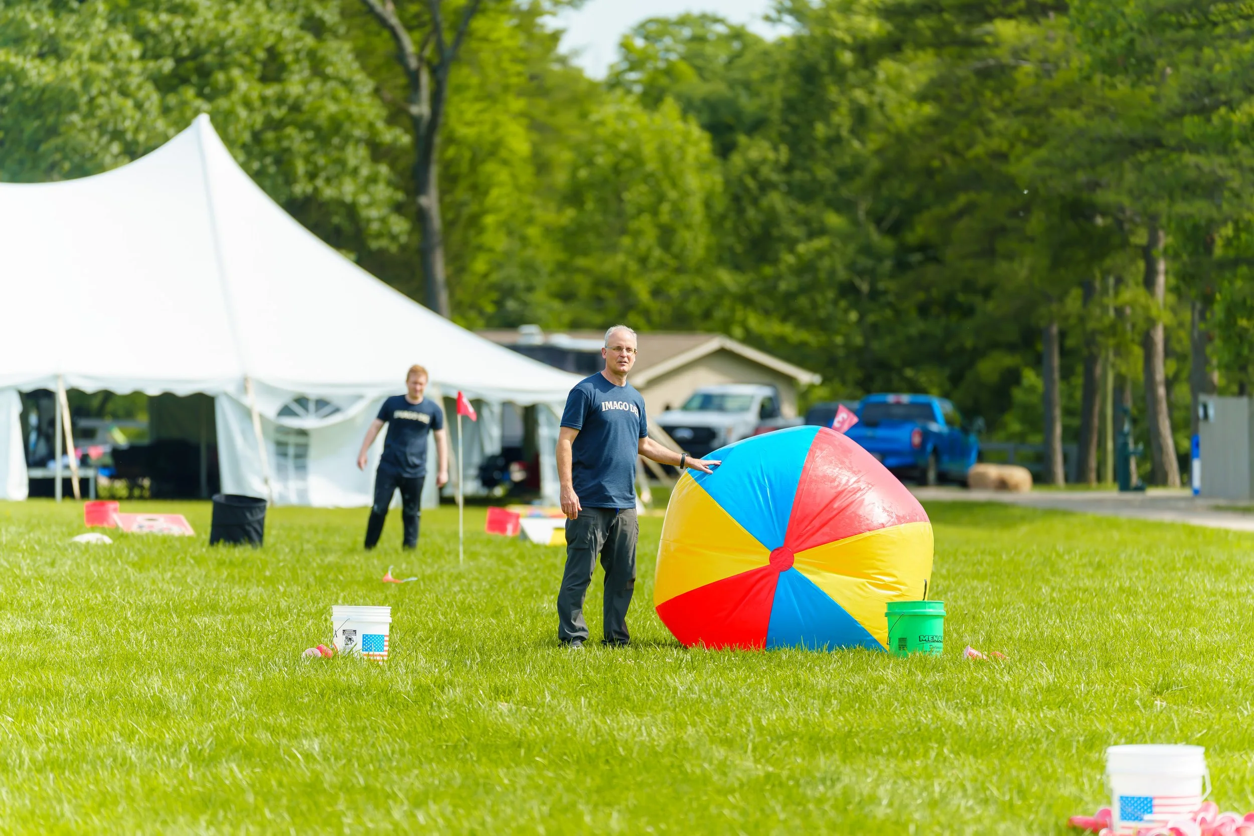 Two men outdoors play with a large colorful beach ball, surrounded by buckets and cones, under a tent with green trees in the background.