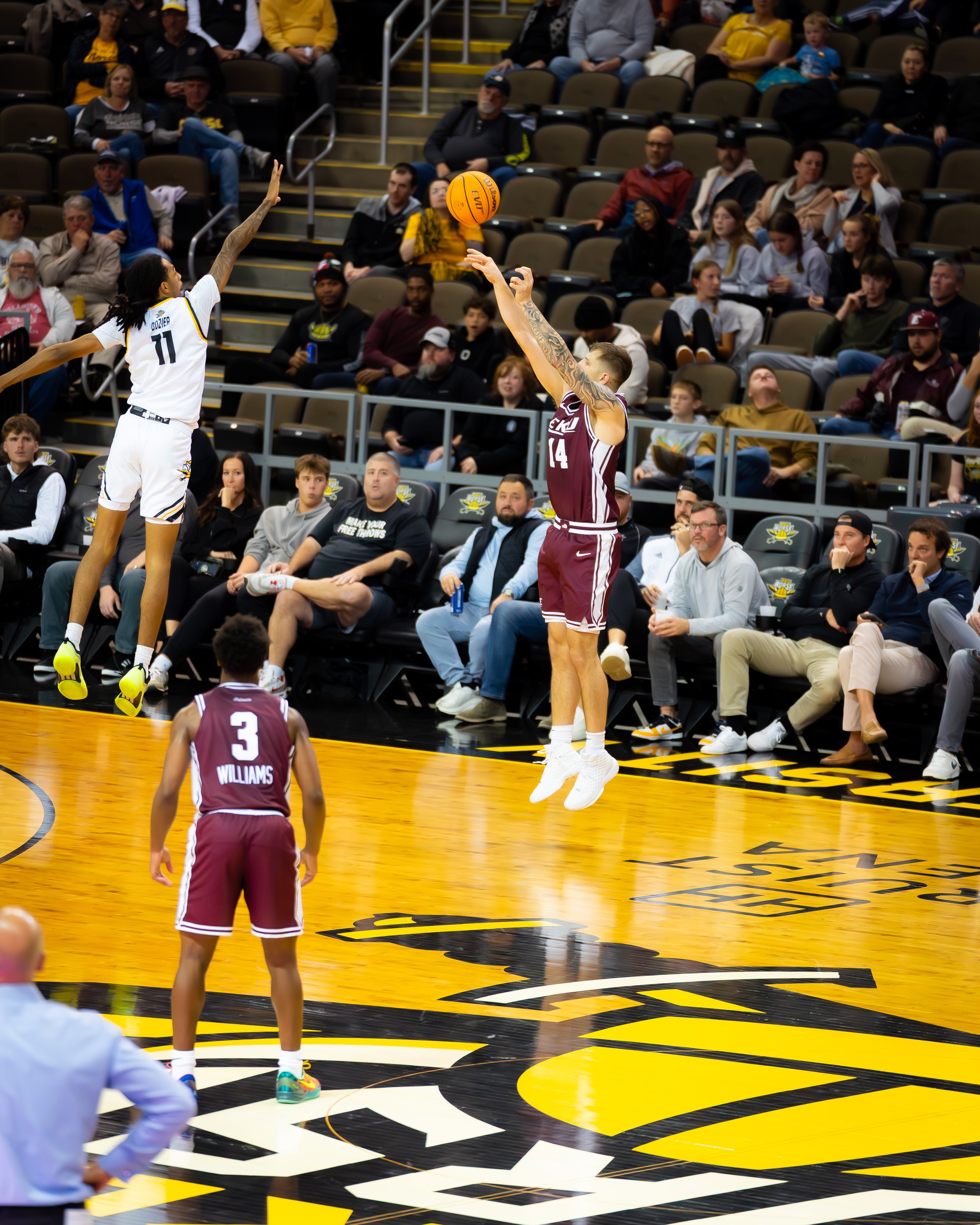 A basketball game with players leaping for the ball in the air, one in a white and yellow uniform and the other in maroon, with spectators sitting in the background.