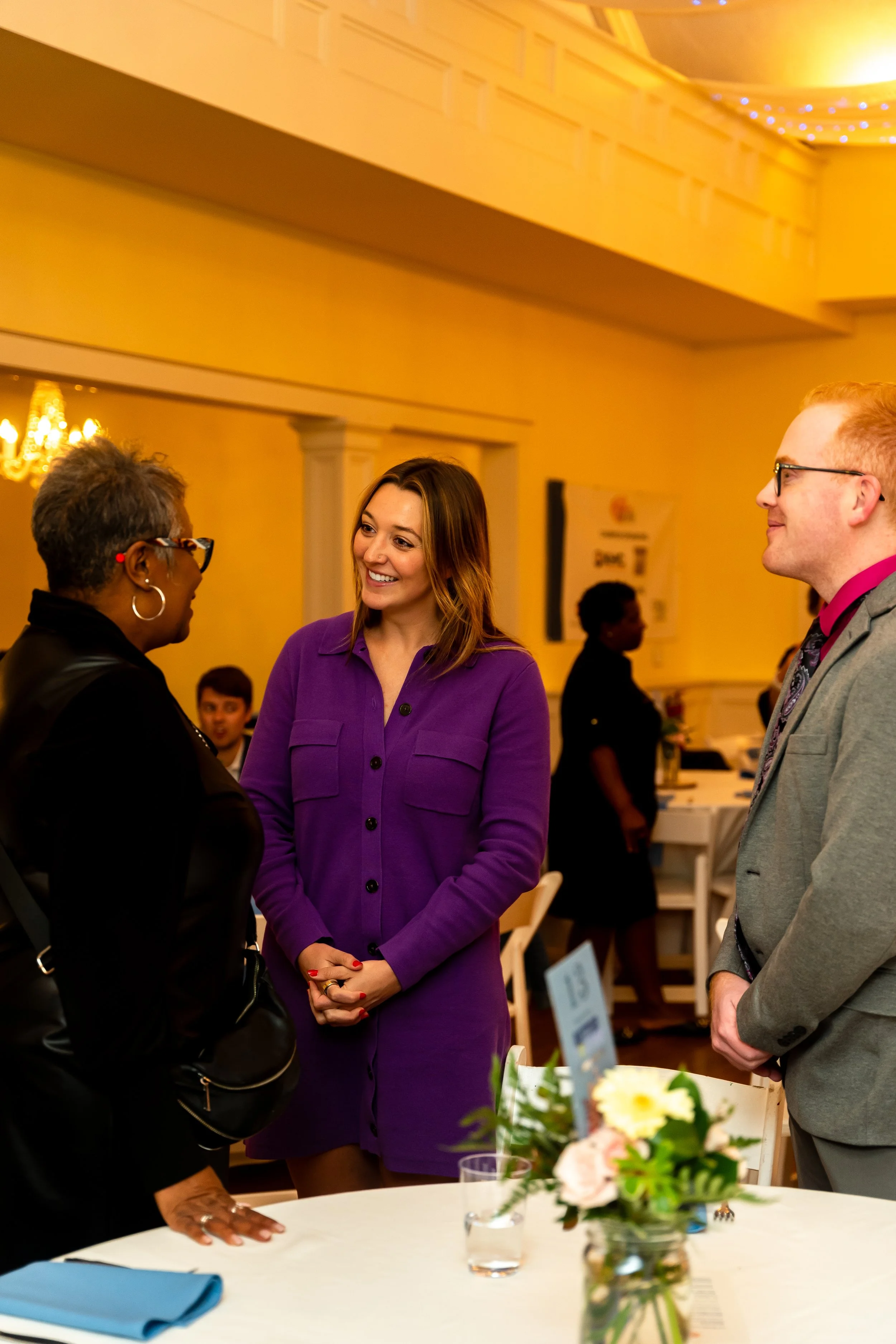 Three people engaged in conversation indoors, with a table in the foreground holding a flower arrangement, a glass of water, and a folded blue napkin. The background shows warm yellow walls, decorative molding, and other individuals at an event.