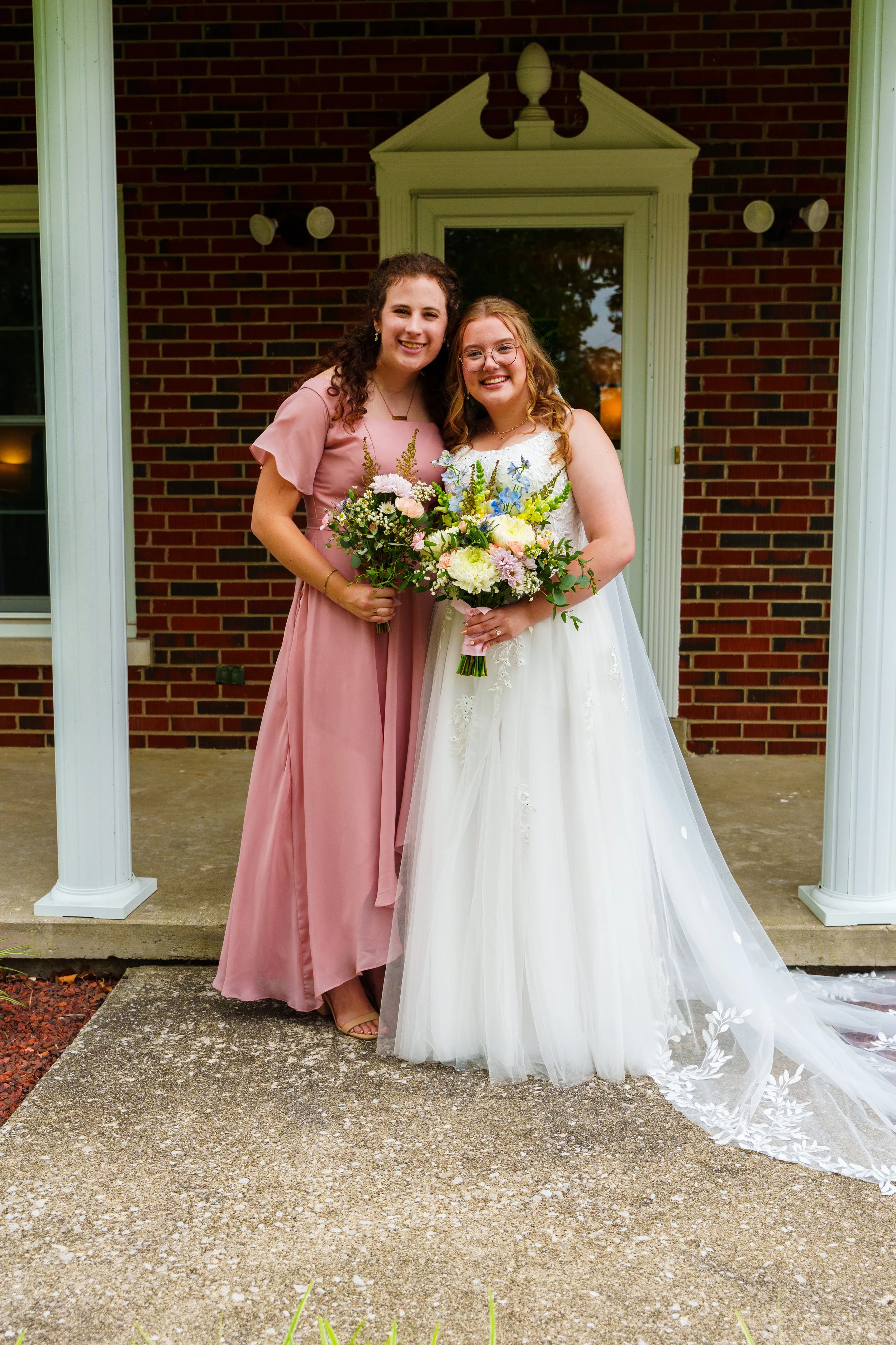 Two women standing outside a house with brick walls and white columns, one in a white wedding dress holding a bouquet of flowers, and the other in a pink dress with a bouquet, smiling together.